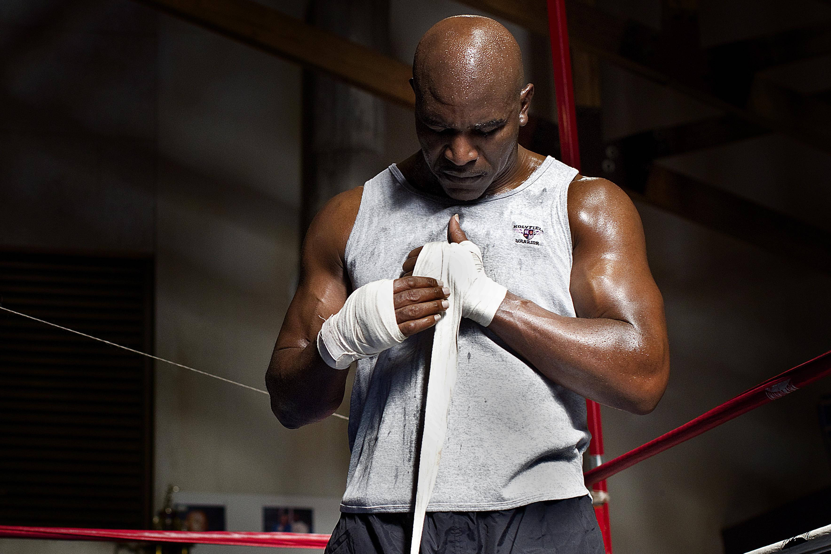 Boxing 桌布 壁紙 — 一個肌肉發達的光頭拳擊手在昏暗的擂台上，在戲劇性的燈光下專注地用白色繃帶纏繞雙手。