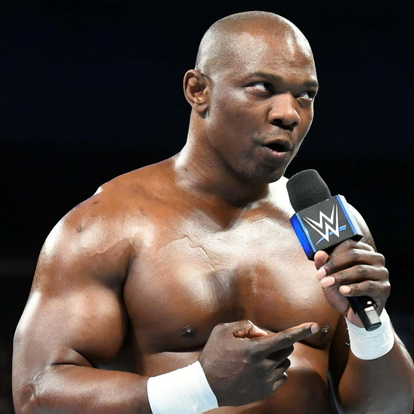 WWE 桌布 壁紙 — 一個肌肉發達的光頭摔角手，戴著白色護腕，在黑暗背景前拿著WWE麥克風並指向前方。