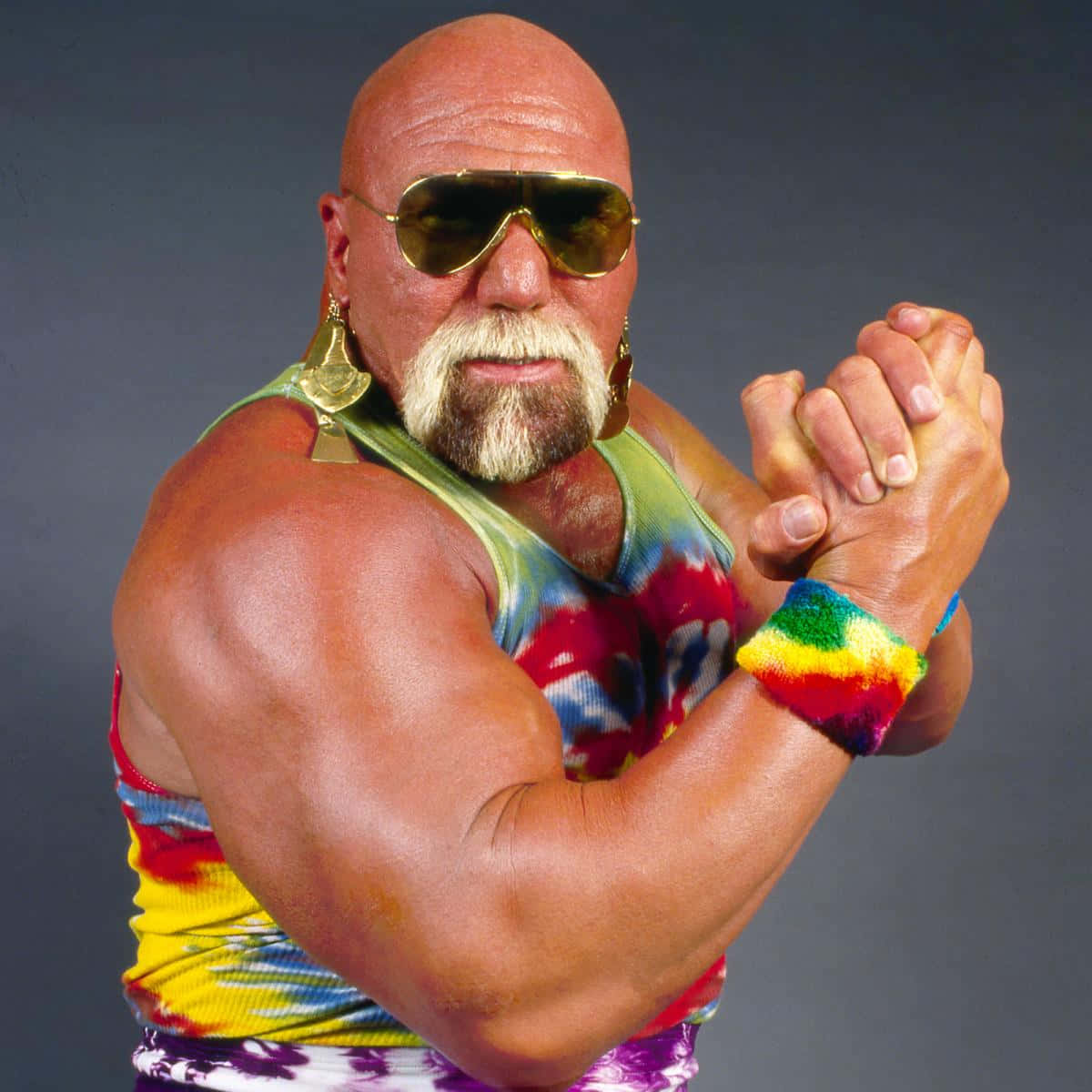 WWE 桌布 壁紙 — 一個肌肉發達的光頭摔角手，留著白色山羊鬍，戴著金色飛行員太陽眼鏡，穿著鮮豔的紮染背心和彩虹腕帶，彎曲手臂。