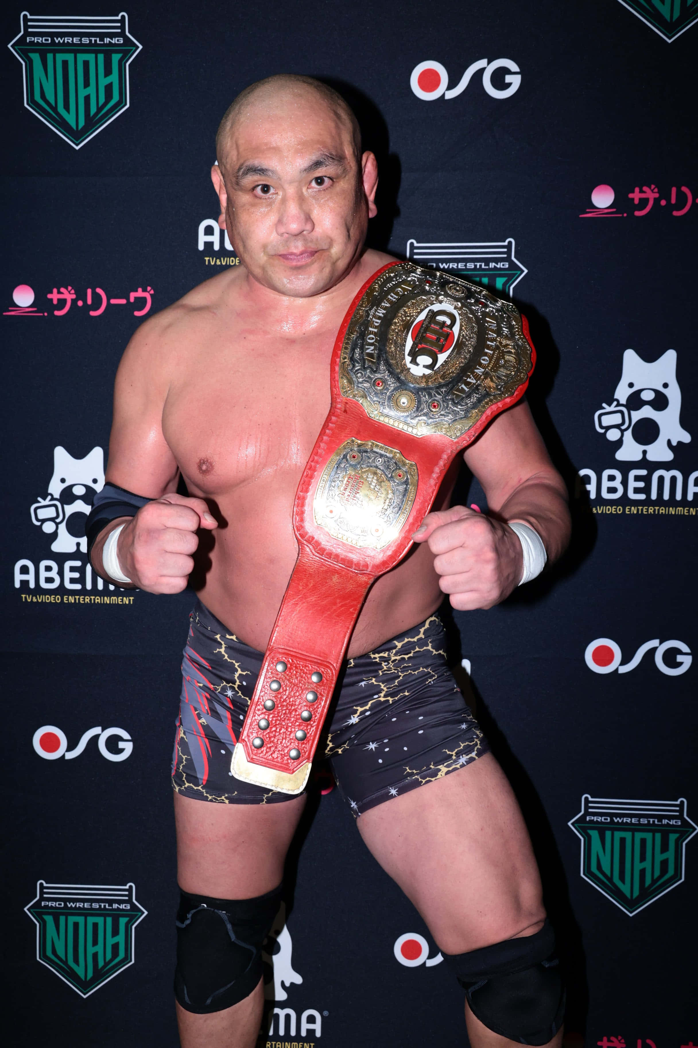 Athlete 桌布 壁紙 — 一個肌肉發達的光頭摔角手自豪地舉著一條紅金色的冠軍腰帶，背景是品牌活動場景。