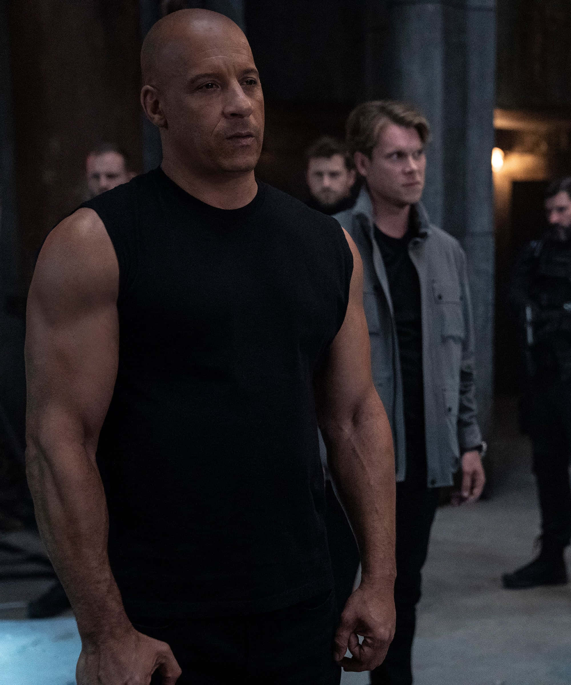 Fast And Furious 桌布 壁紙 — 一個肌肉發達的光頭男人穿著黑色背心，緊張地站在光線昏暗的工業走廊裡，身後有其他男人。