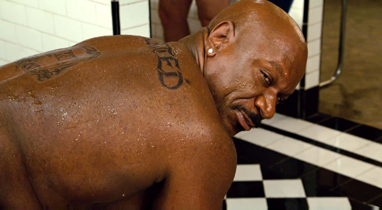 Ving Rhames 桌布 壁紙 — 一個肌肉發達的光頭男子，身上有刺青和戴著耳環，在磁磚浴室裡向前傾，水滴在他皮膚上閃閃發光。
