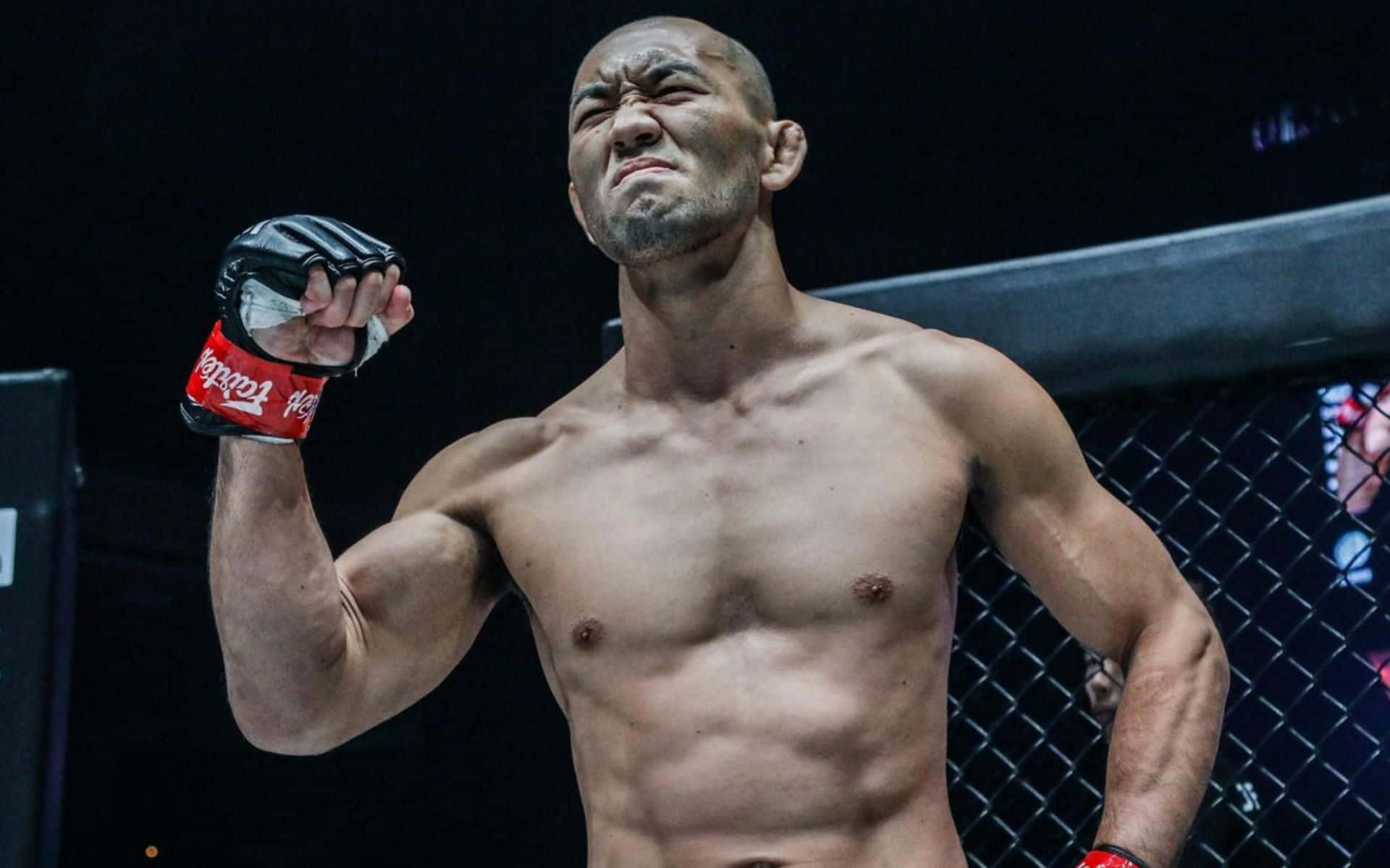 UFC 桌布 壁紙 — 一個肌肉發達的拳手在籠中高舉戴著手套的拳頭，面露兇猛表情，在明亮的場館燈光下慶祝勝利。