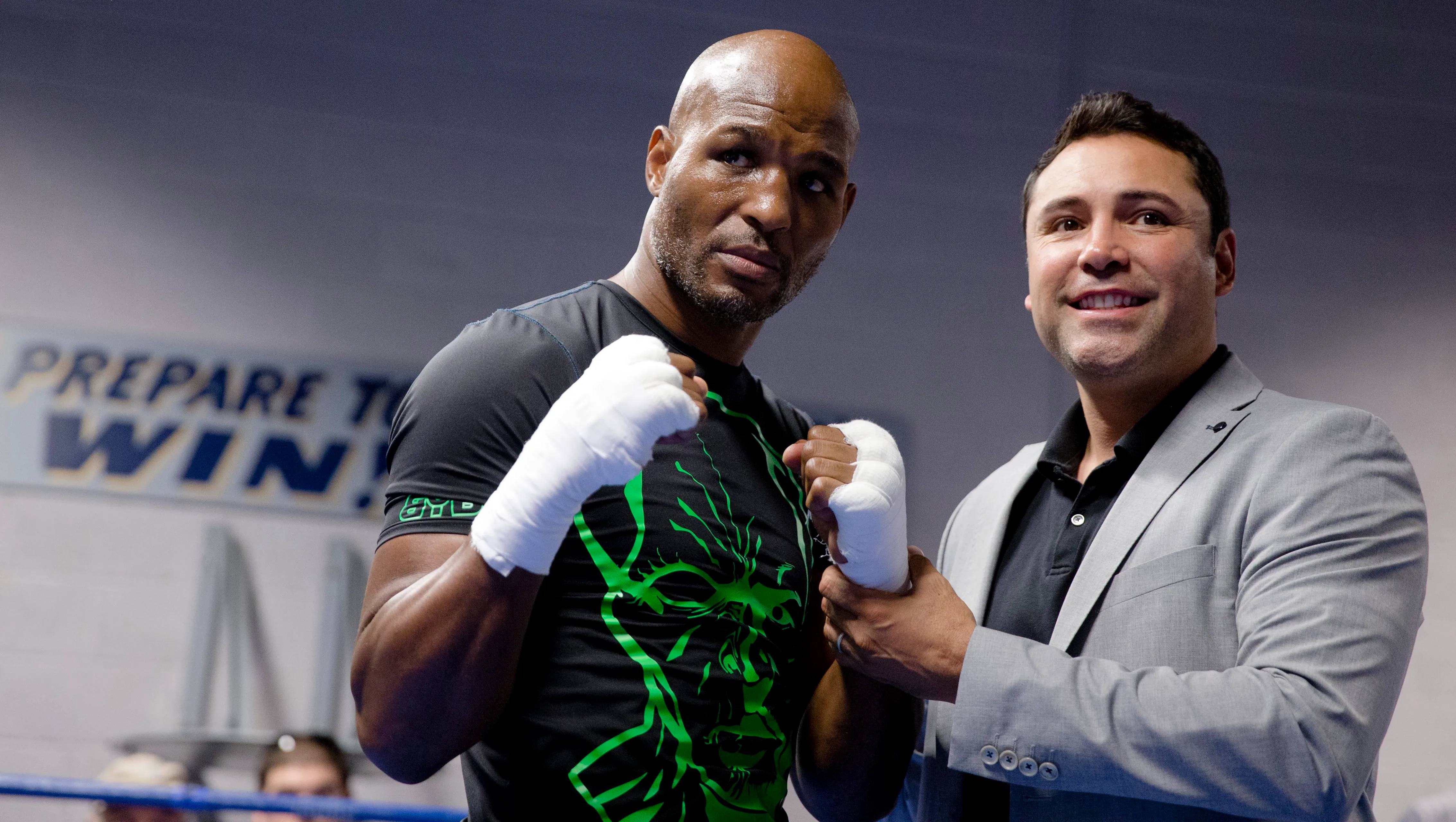 Boxing 桌布 壁紙 — 一個肌肉發達的拳擊手穿著帶有綠色骷髏圖案的黑色襯衫，戴著白色手套站在擂台上，一名穿著灰色西裝的男子正在為他纏手。