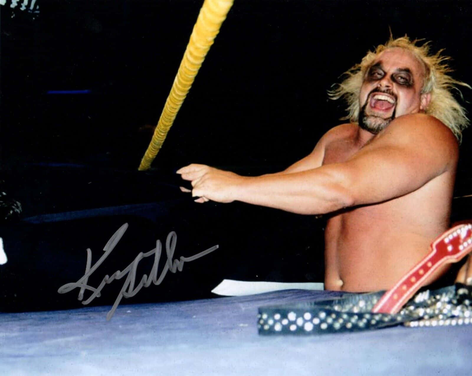 WWE 桌布 壁紙 — 一個肌肉發達的摔角手，有著狂野的金髮和黑色臉部塗料，在擂台上尖叫，手持吉他，附近有一條鉚釘腰帶。
