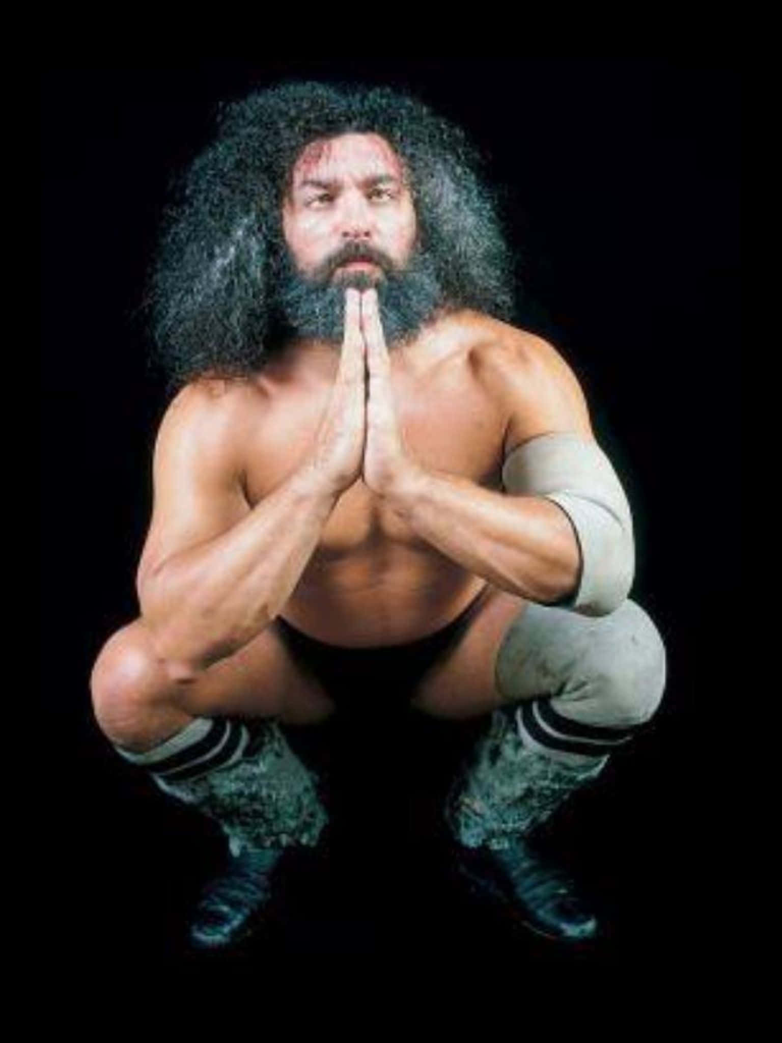 WWE 桌布 壁紙 — 一個肌肉發達的摔角手，留著狂野鬍鬚和長頭髮，在黑色背景前蹲低祈禱。