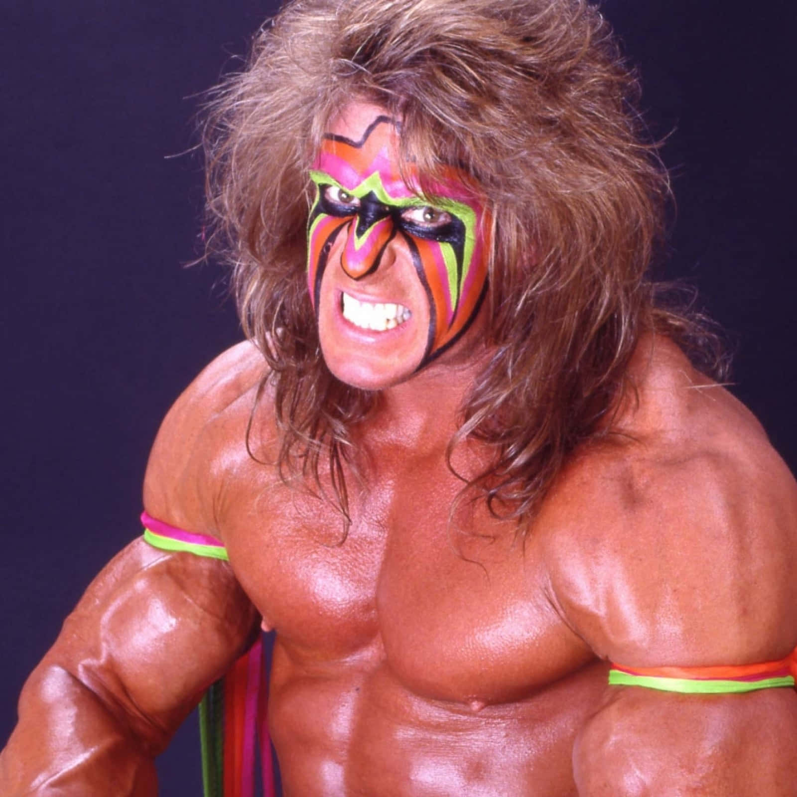 WWE 桌布 壁紙 — 一個肌肉發達的摔角手，頭髮狂野，臉上塗著鮮豔的顏料，在黑暗的背景下做出強烈的鬼臉。