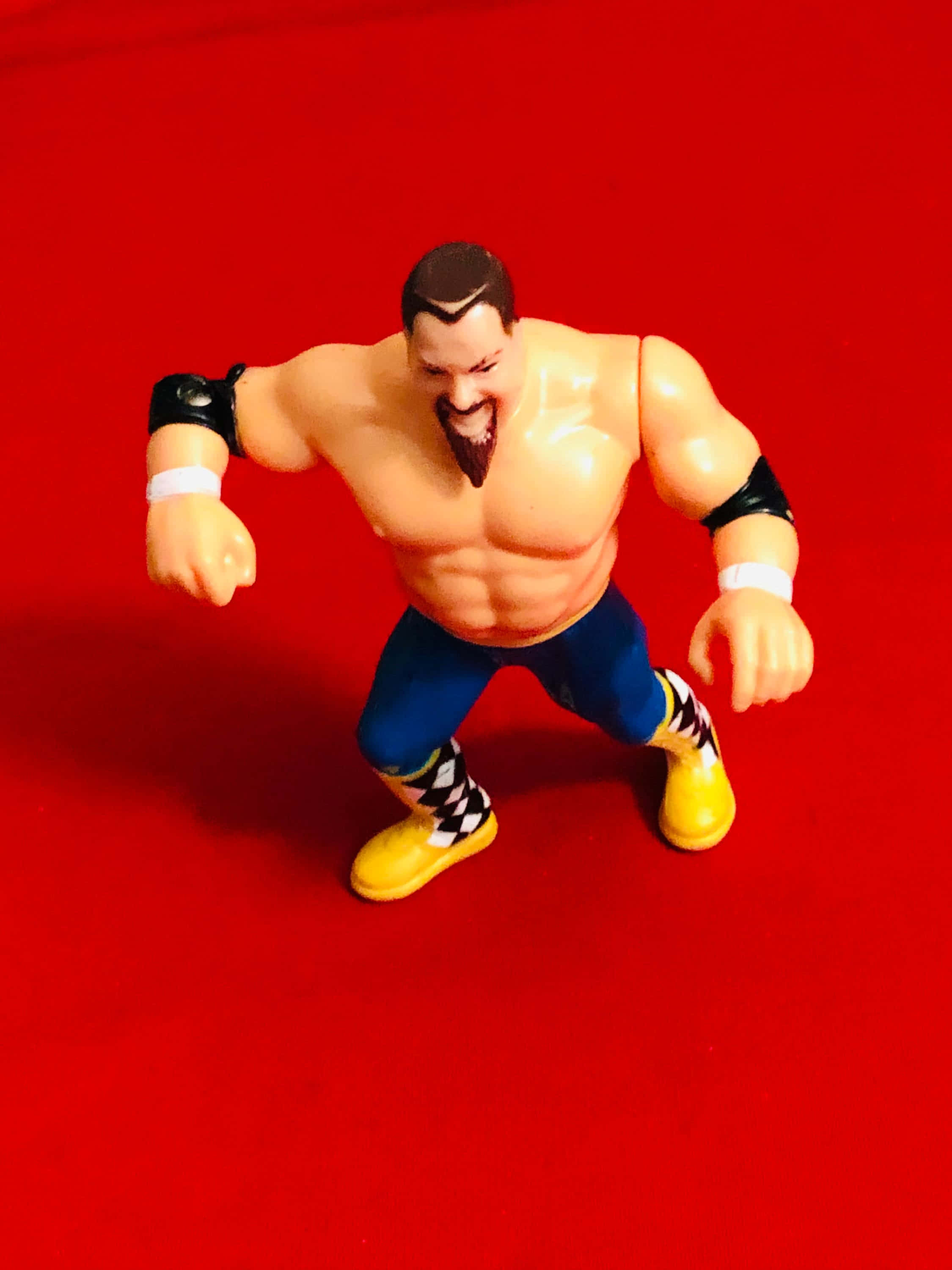 Wrestling 桌布 壁紙 — 一個肌肉發達的摔角手動作人偶，穿著藍色短褲和黃色靴子，在純紅色背景上擺出挑釁姿勢。