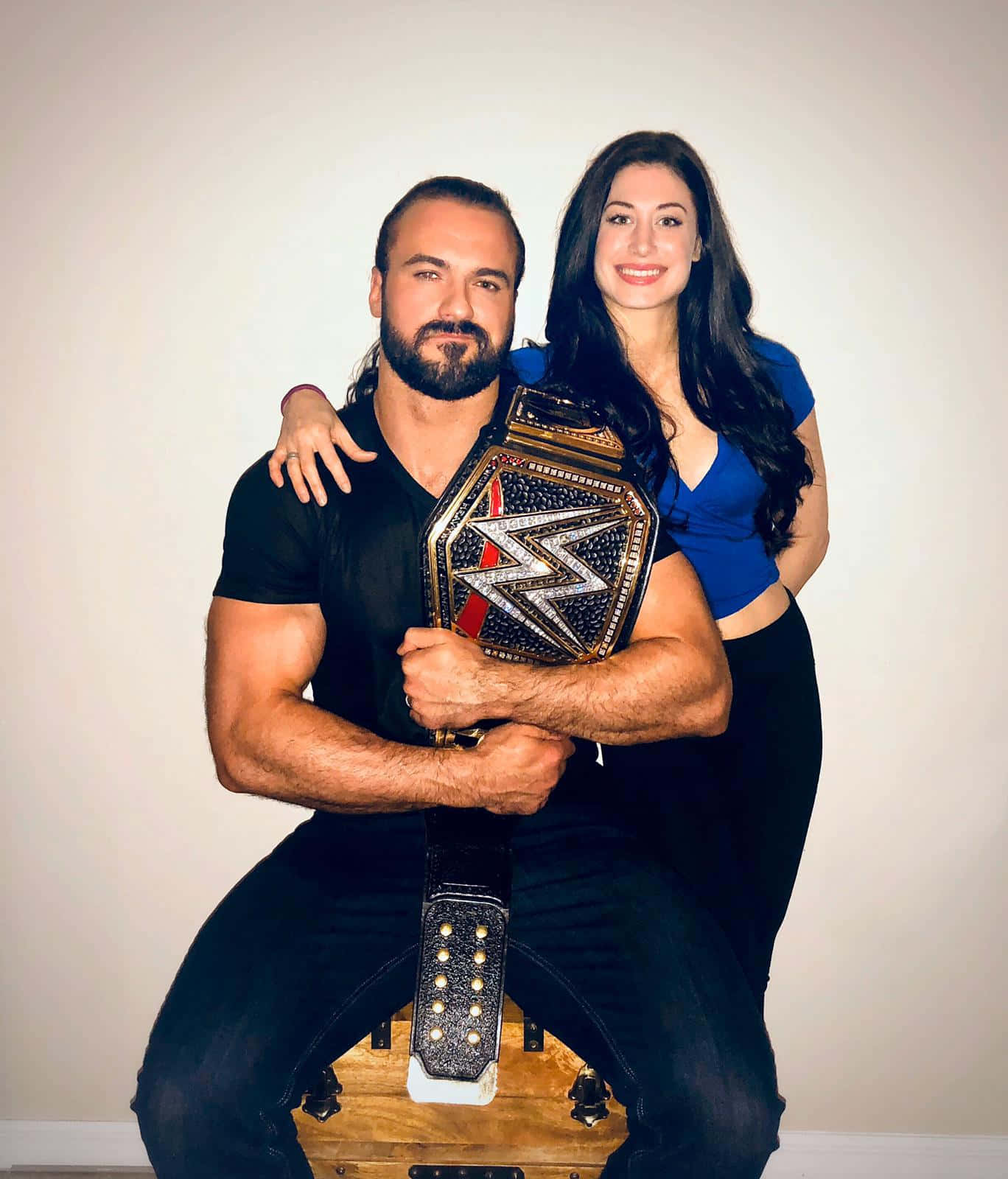 WWE 桌布 壁紙 — 一個肌肉發達的摔角手穿著黑色襯衫，手持WWE冠軍腰帶，而一個微笑的女人穿著藍色上衣從後面擁抱他，背景是素牆。