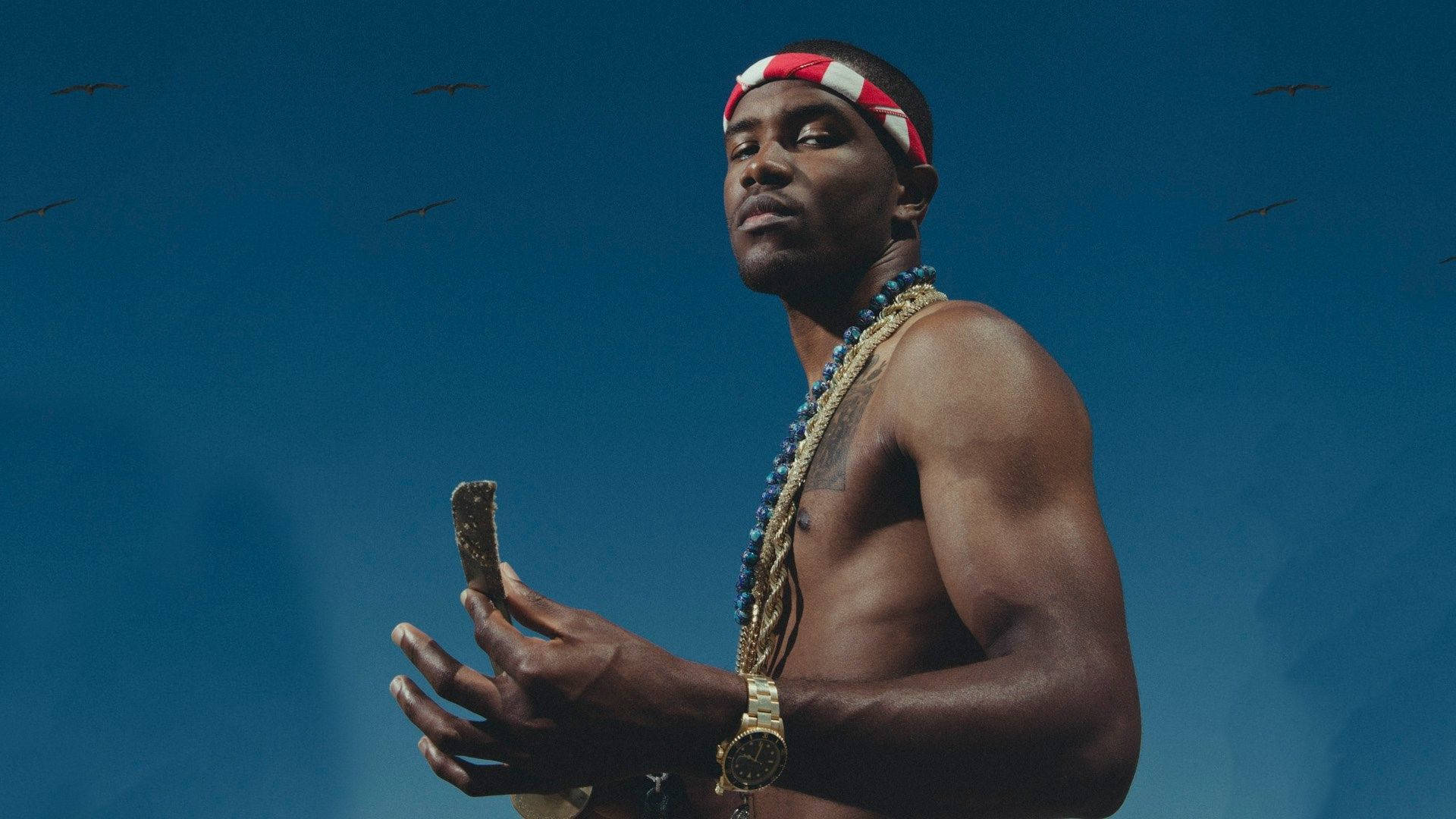 Frank Ocean 桌布 壁紙 — 一個肌肉發達的男人，戴著紅白頭帶和金鏈，站在深藍色的天空下，鳥兒在頭頂飛翔。