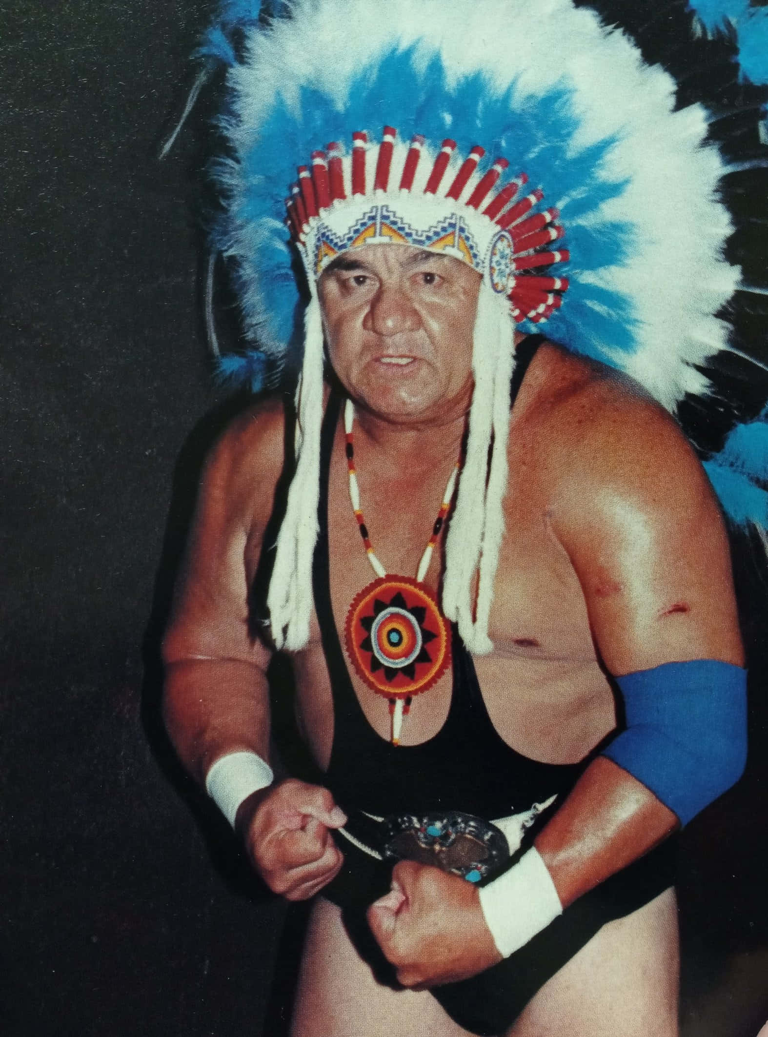 Native American 桌布 壁紙 — 一個肌肉發達的男人，戴著藍白羽毛頭飾和部落項鍊，在黑暗背景下強烈地擺姿勢。