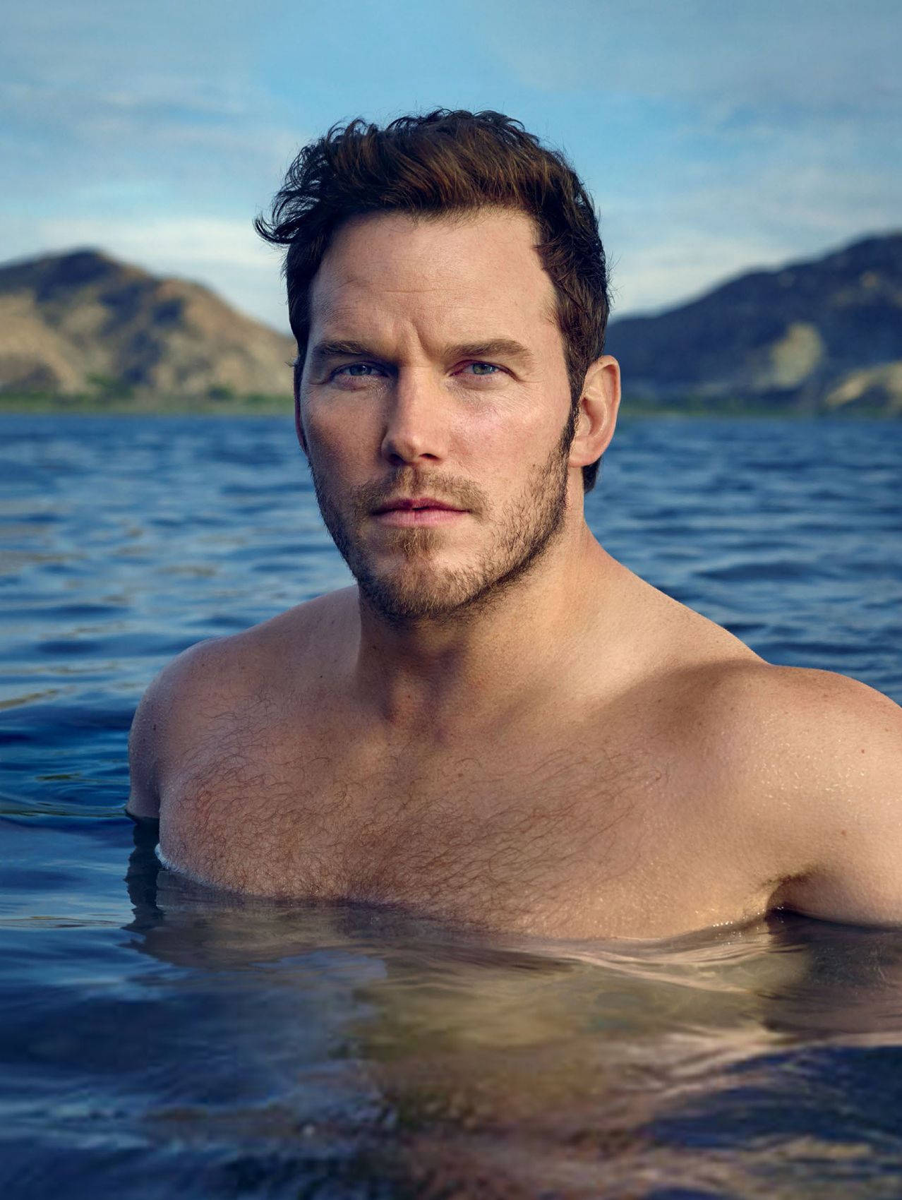 Chris Pratt 桌布 壁紙 — 一個肌肉發達的男人，濕髮和鬍鬚，從平靜的藍色湖泊中凝視著遠方的山丘，天空晴朗。