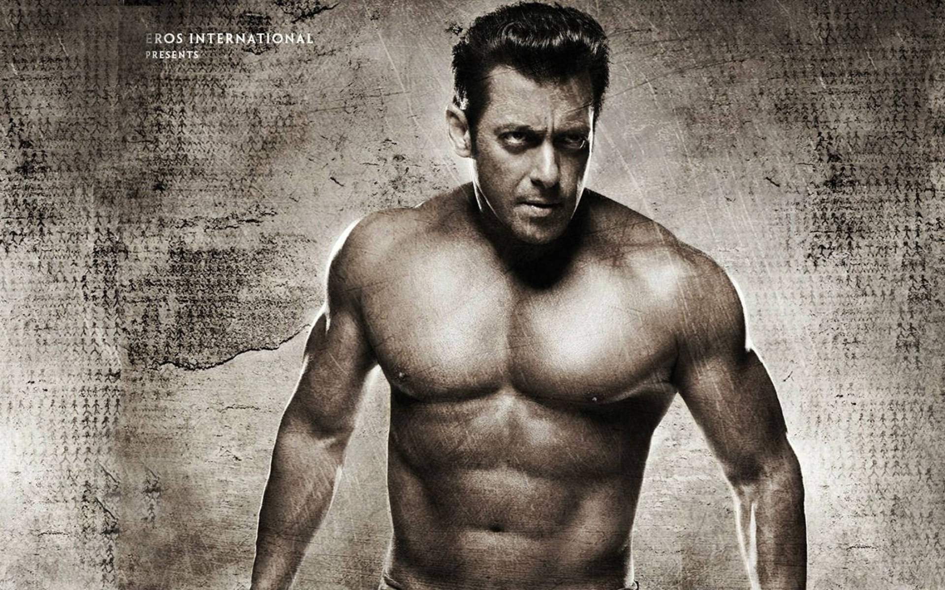 Salman Khan 桌布 壁紙 — 一個肌肉發達的男人，目光銳利，赤裸上身站在有紋理的粗糙牆壁前，在戲劇性的黑白燈光下。