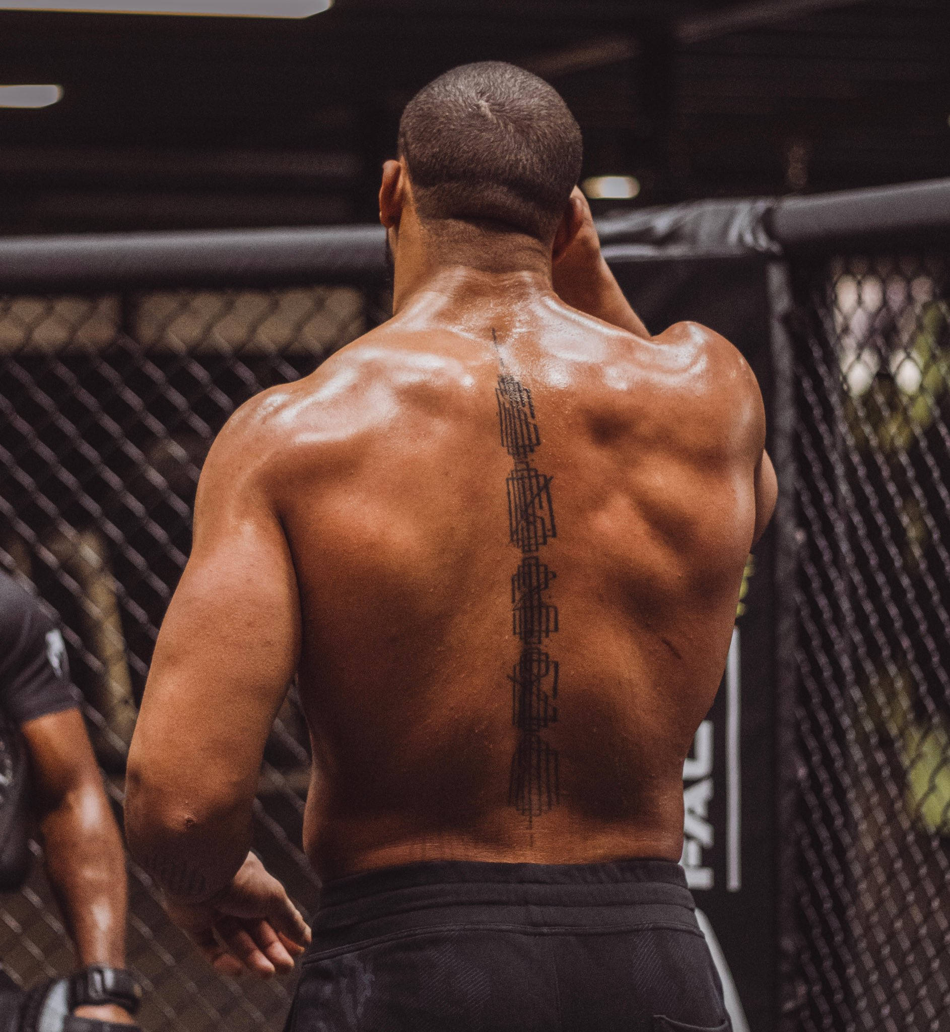 UFC 桌布 壁紙 — 一個肌肉發達的男人，背部有垂直刺青，站在MMA八角籠內，汗水在背上閃閃發光。