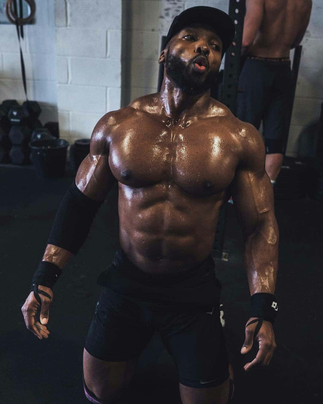 Muscle 桌布 壁紙 — 一個肌肉發達的男人，閃亮的上半身和鬍鬚，站在健身房，戴著帽子和手腕帶，向上看，充滿強烈的眼神。