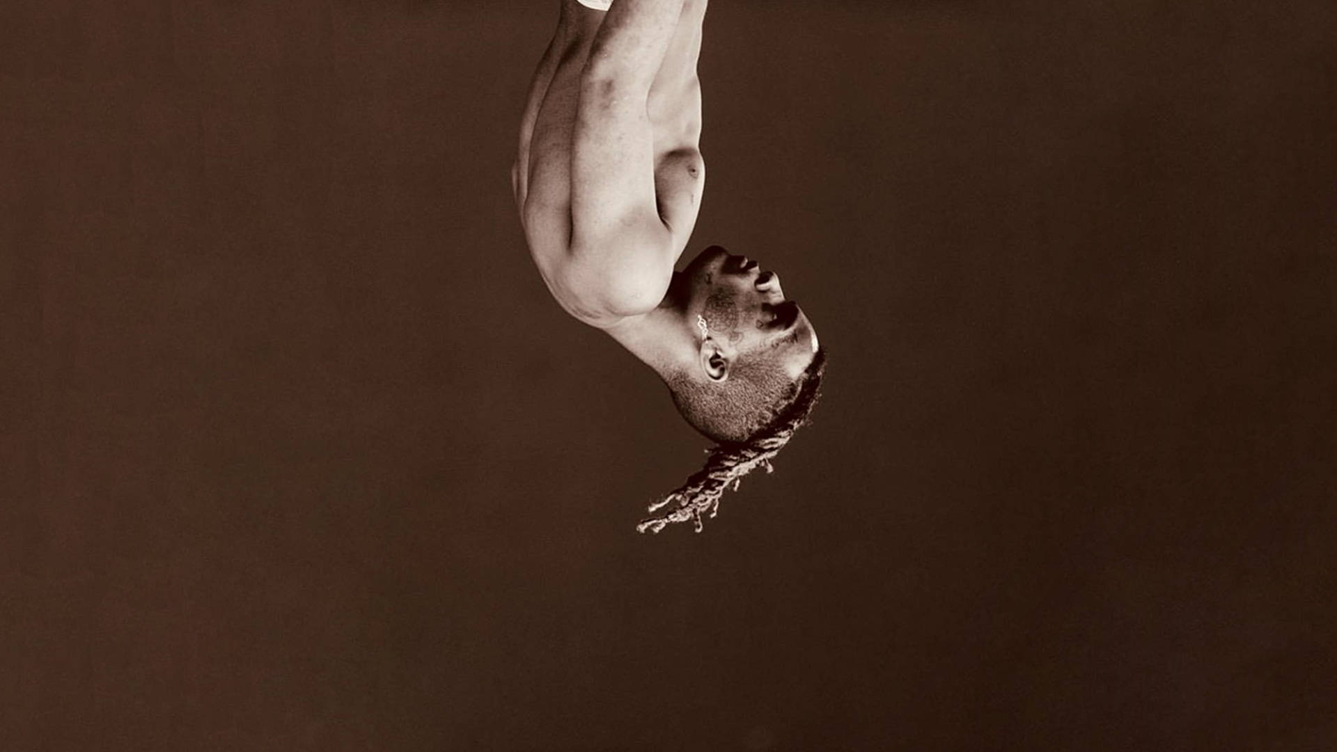 xxxtentacion 電腦 桌布 壁紙 — 一個肌肉發達的男人倒掛在柔和的棕色背景前，展示出清晰的雙臂和辮子，營造出戲劇性的攝影棚效果。