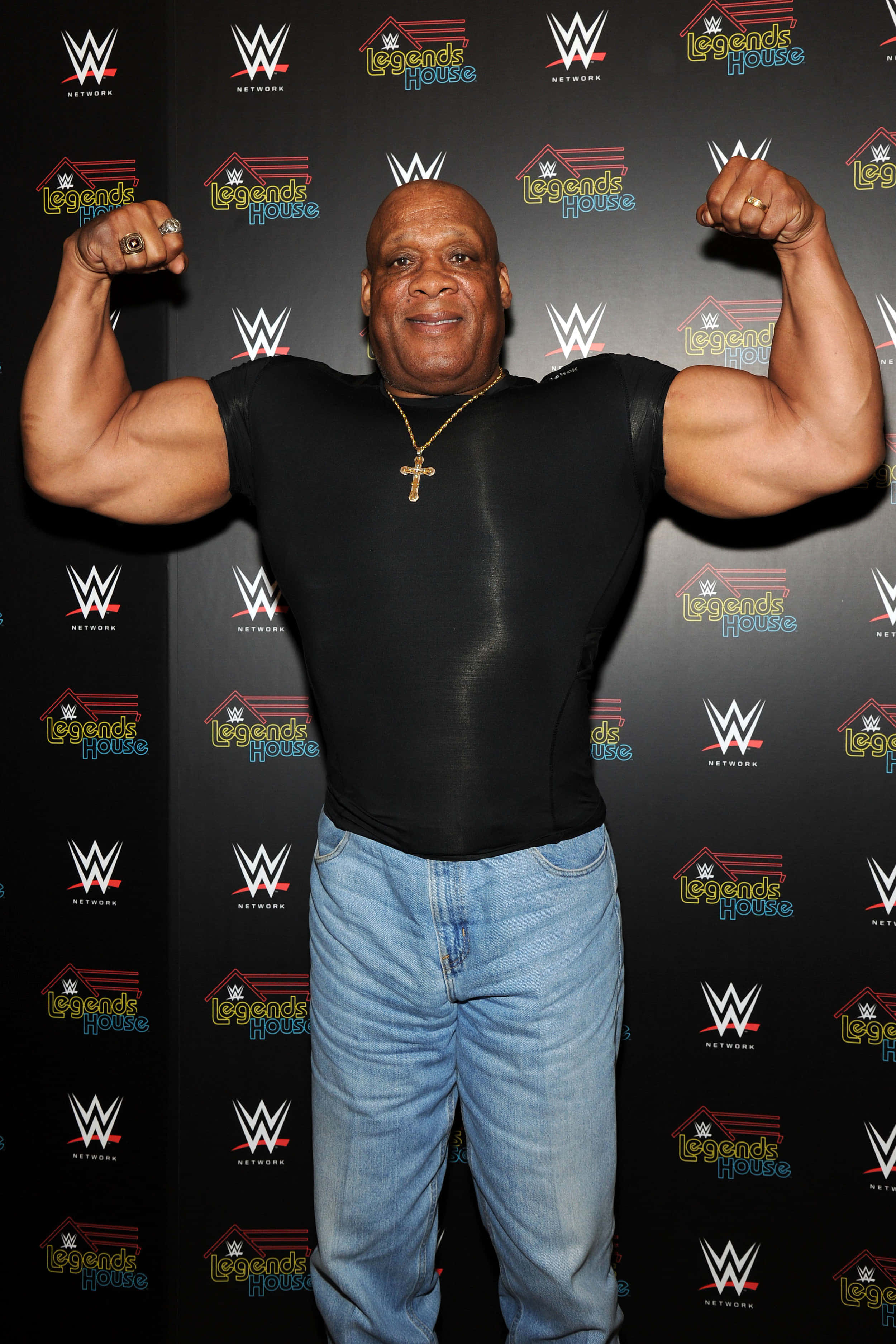 摔角 桌布 壁紙 — 一個肌肉發達的男人在WWE Legends House品牌背景前彎曲他的二頭肌，穿著黑色襯衫和牛仔褲。