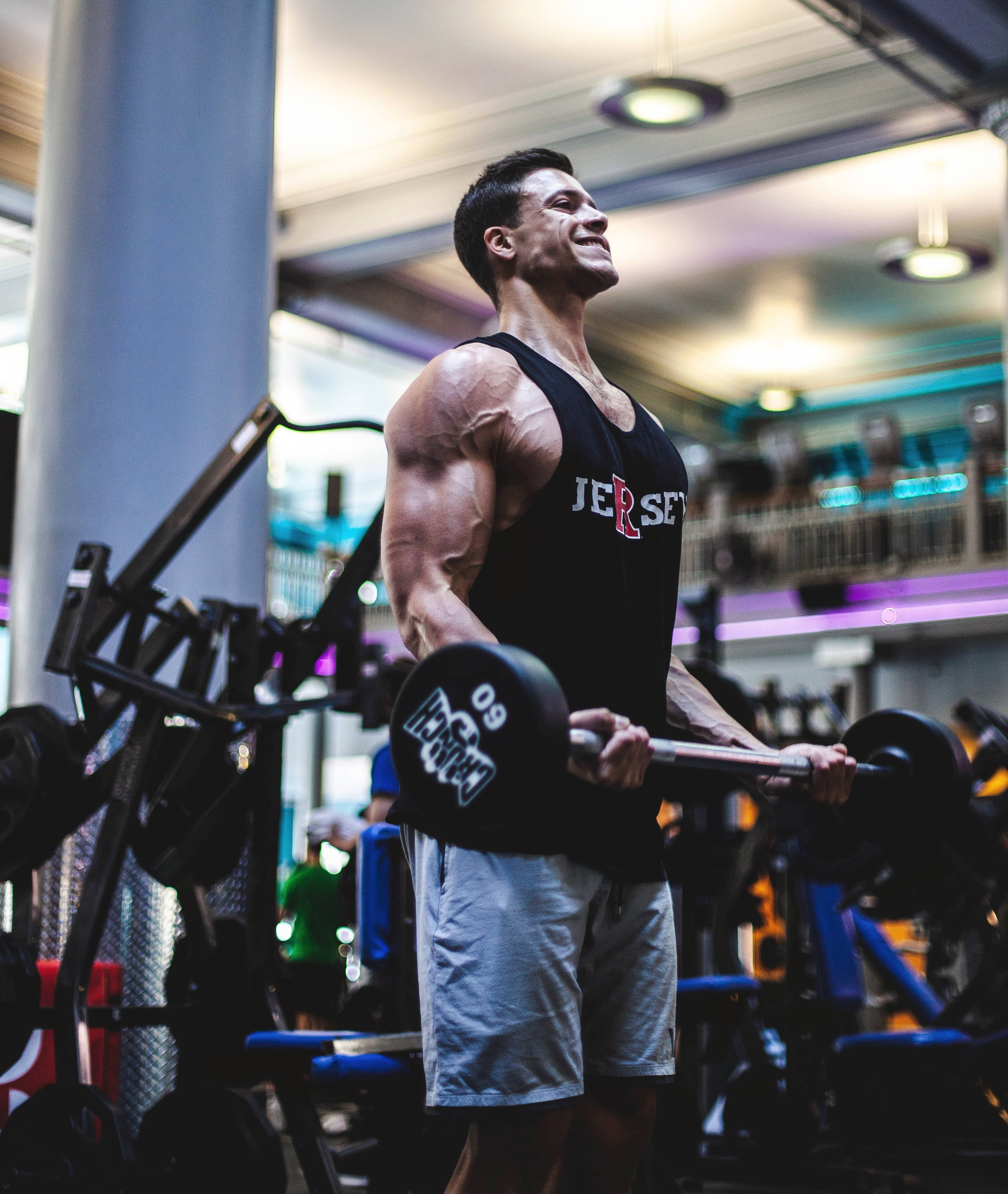 Barbell 桌布 壁紙 — 一個肌肉發達的男人在光線明亮的健身房裡微笑著舉起槓鈴，周圍是色彩繽紛的器材。