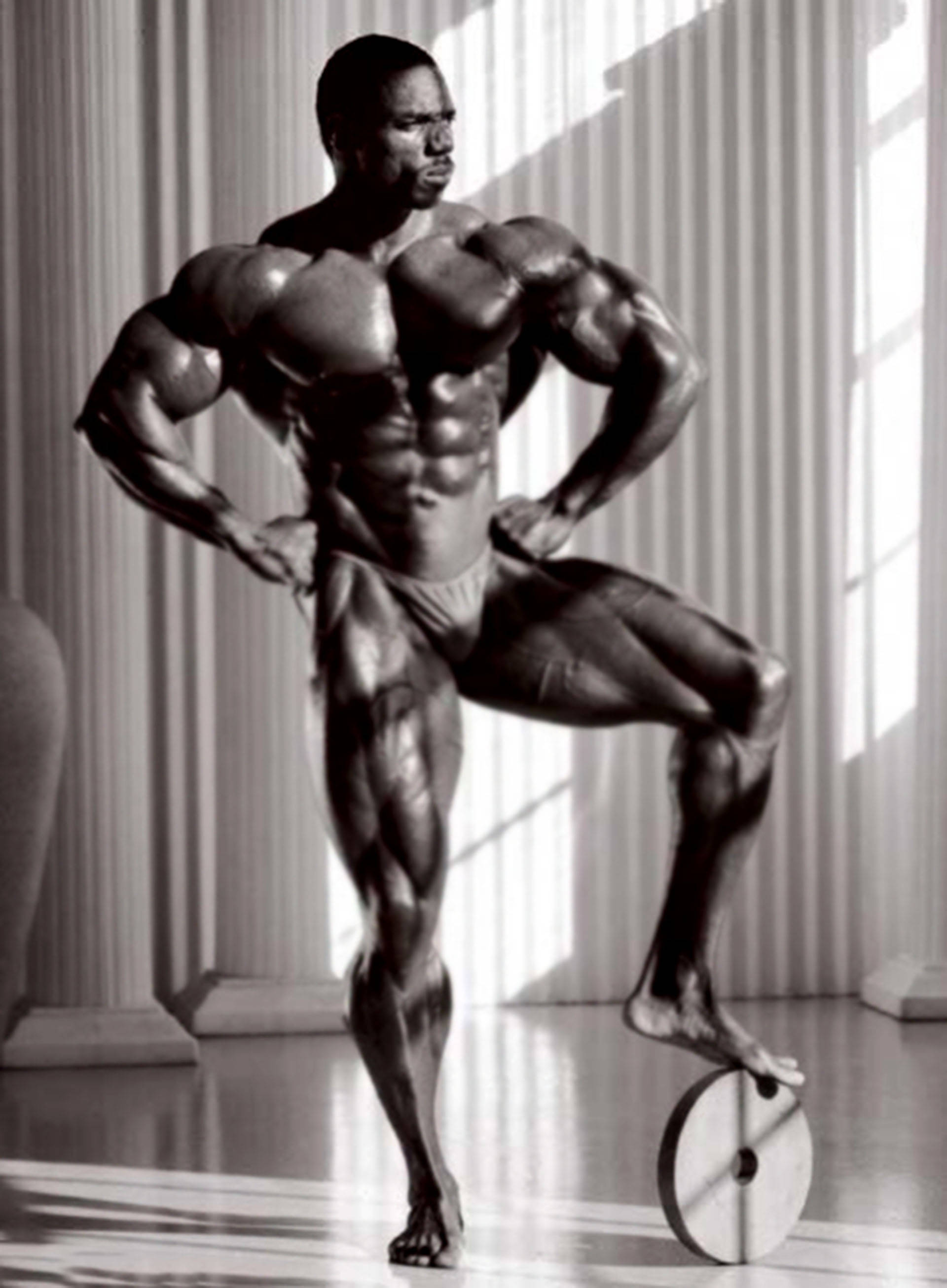 flex wheeler 桌布 壁紙 — 一個肌肉發達的男人在單色調的工作室中，帶著戲劇性燈光和古典柱，自信地擺姿勢。