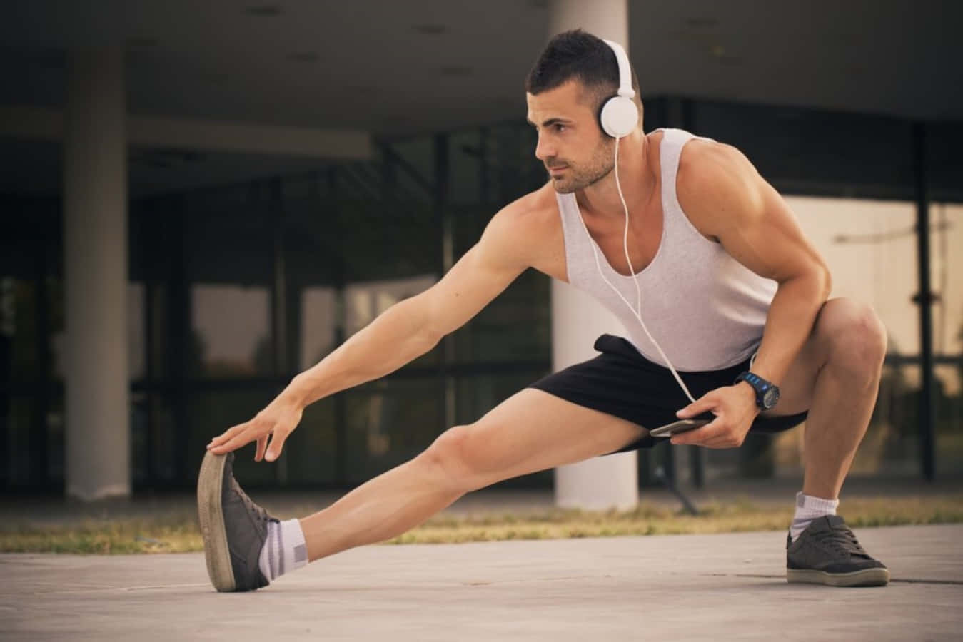 Fitness 桌布 壁紙 — 一個肌肉發達的男人在戶外伸展，戴著白色耳機，拿著電話，蹲在現代建築附近的人行道上。