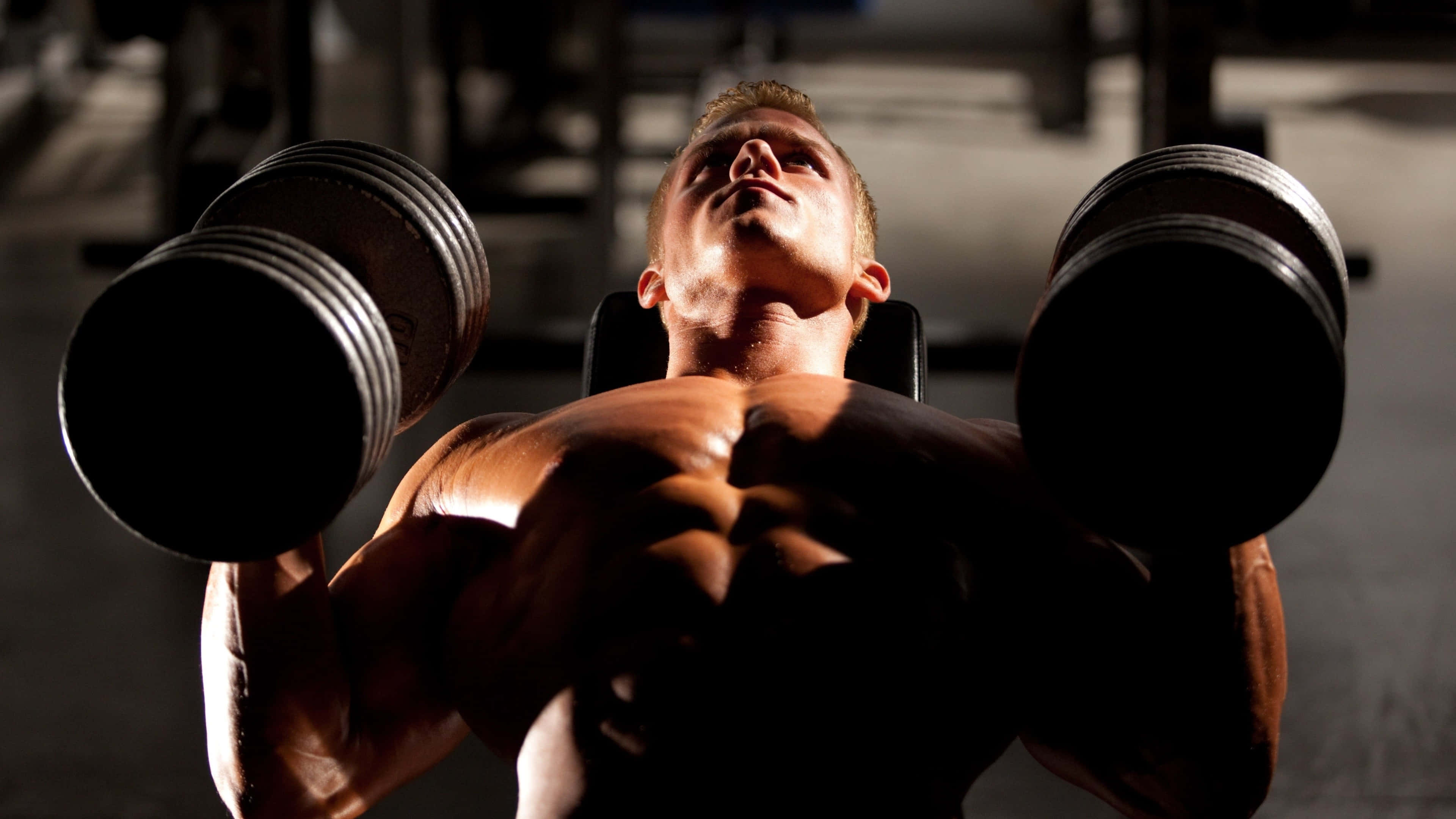 Fitness 桌布 壁紙 — 一個肌肉發達的男人在昏暗的健身房中，專注地舉起沉重的啞鈴。
