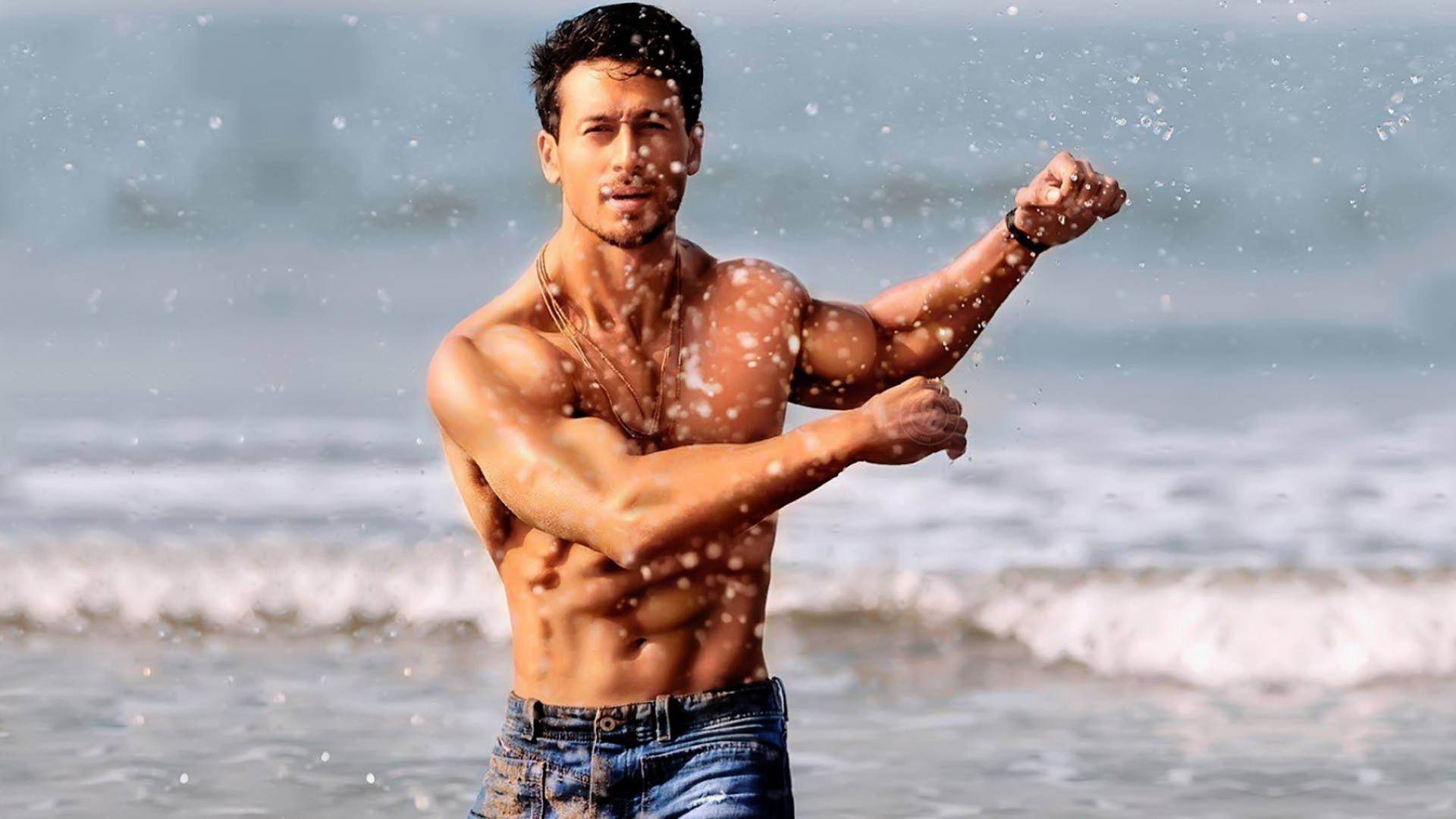 Indian 桌布 壁紙 — 一個肌肉發達的男人在海灘上彎曲手臂，海浪飛濺在明亮的日光中圍繞著他。