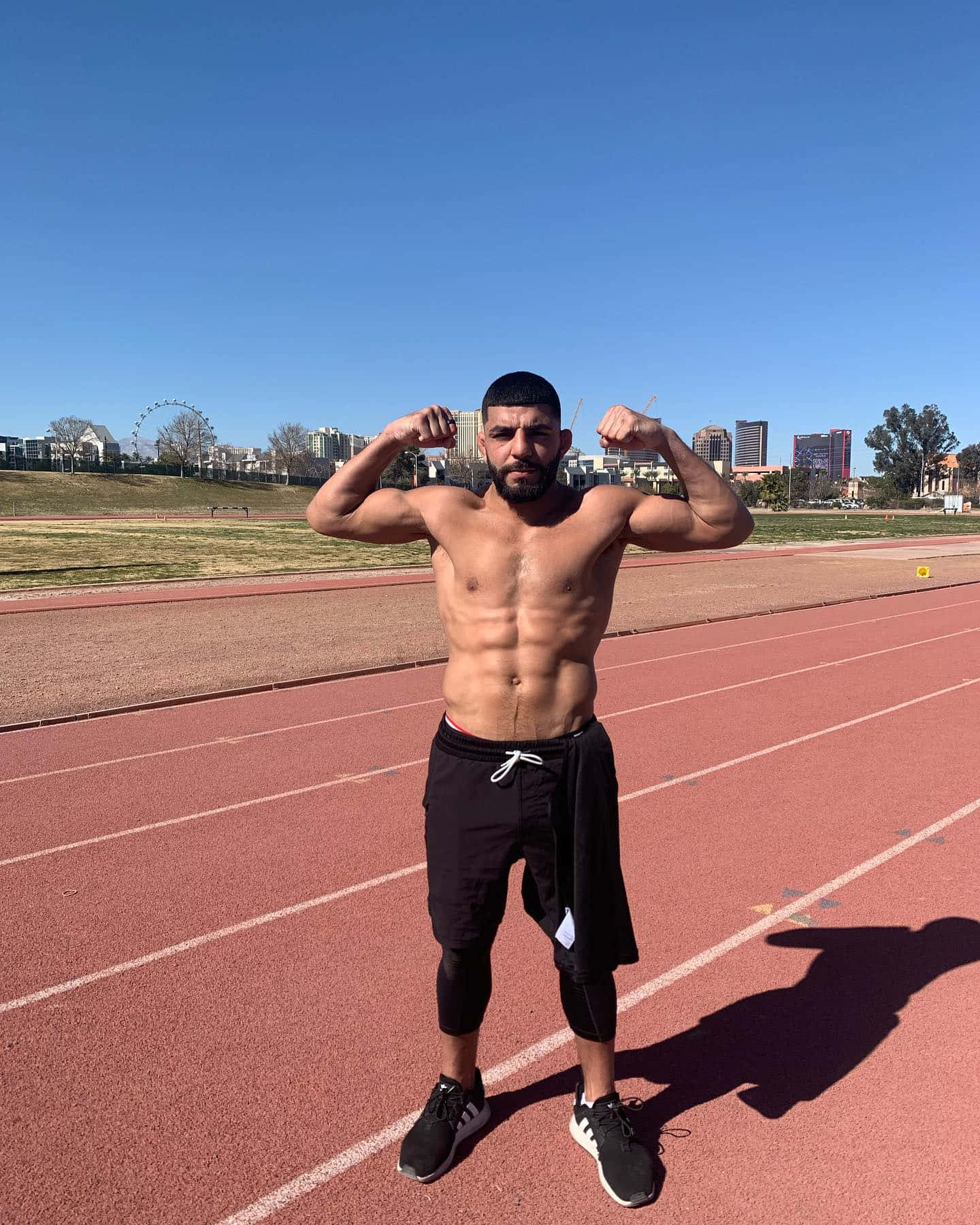 UFC 桌布 壁紙 — 一個肌肉發達的男人在紅色跑道上彎曲手臂，背景是晴朗藍天和城市建築。