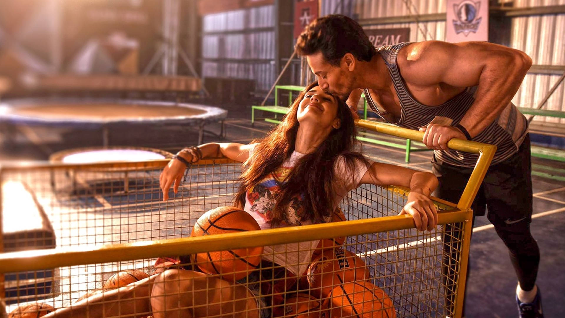 Tiger Shroff 桌布 壁紙 — 一個肌肉發達的男人在陽光普照的健身房裡，親吻一個靠在裝滿球的籃球框上的女人。