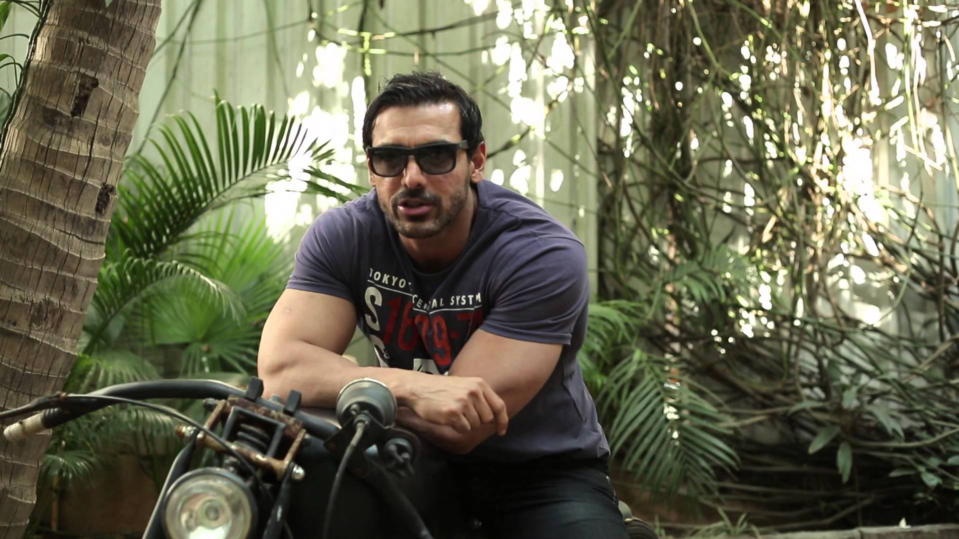 Bollywood HD 桌布 壁紙 — 一個肌肉發達的男人戴著太陽眼鏡，靠在一輛復古機車上，周圍是茂密的熱帶植物，斑駁的陽光透過樹葉灑落。