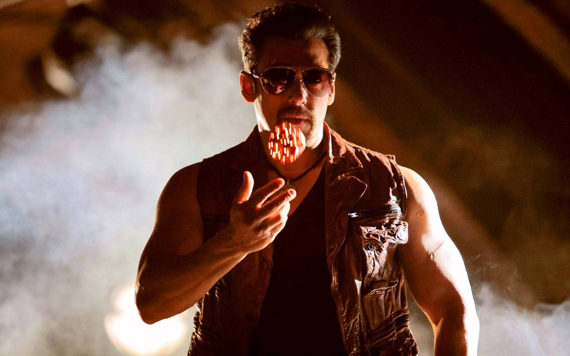 Salman Khan 桌布 壁紙 — 一個肌肉發達的男人戴著太陽眼鏡和背心，手持一個發光的紅色物體，煙霧在他周圍旋轉，環境昏暗。