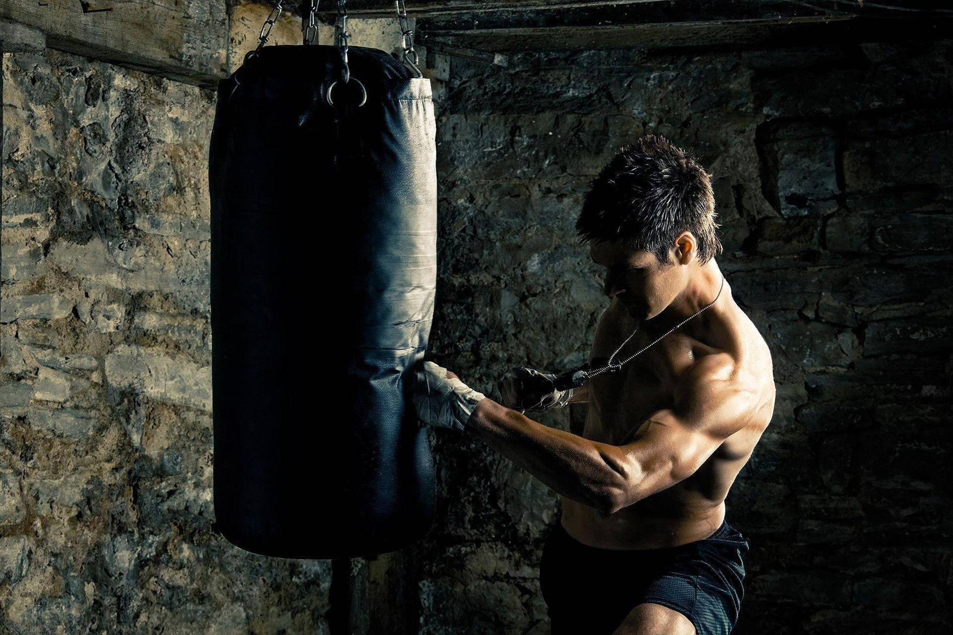 MMA 桌布 壁紙 — 一個肌肉發達的男人戴著手套在光線昏暗的石牆健身房中，以戲劇性的燈光重擊沙包。