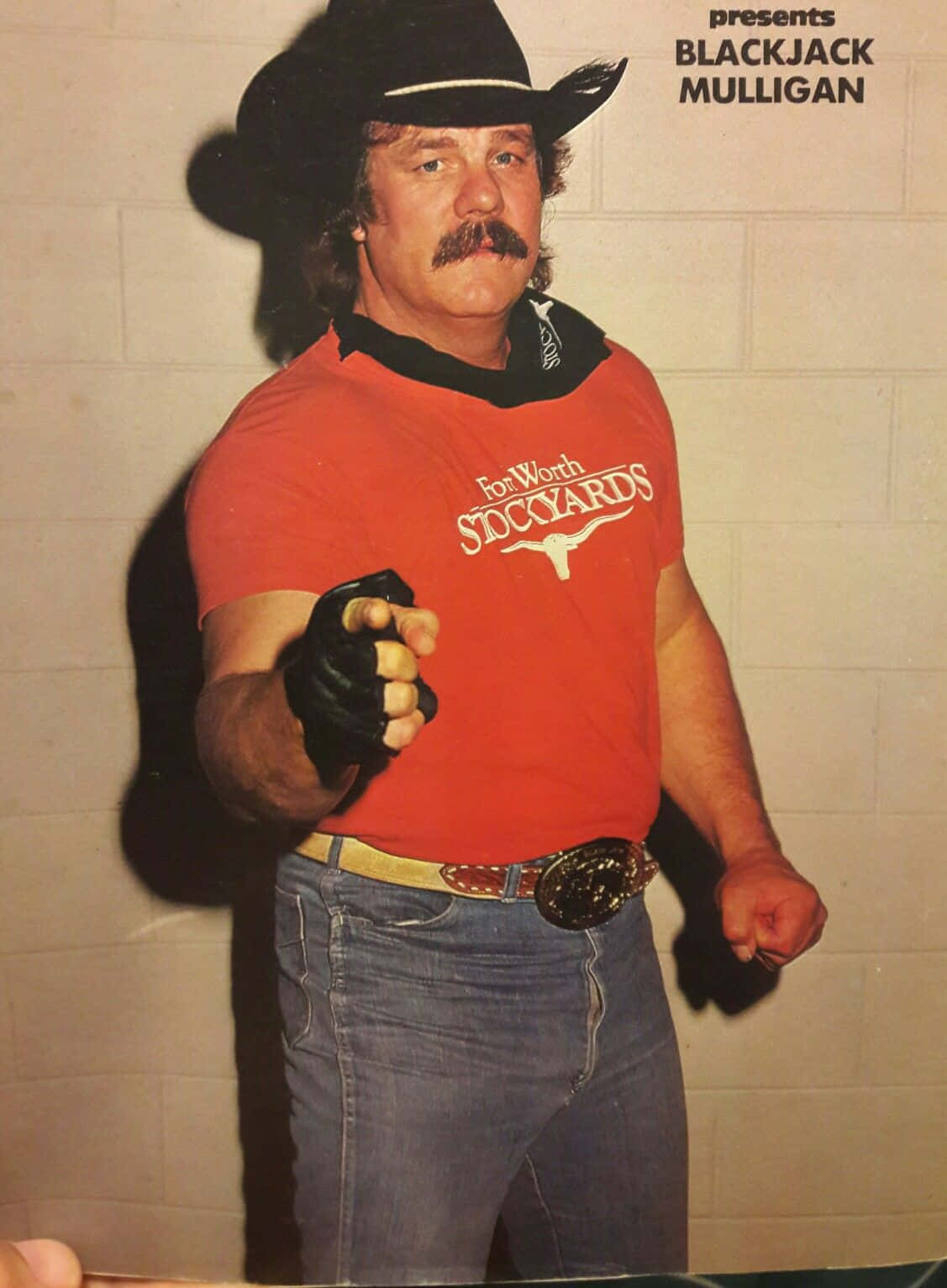 WWE 桌布 壁紙 — 一個肌肉發達的男人戴著牛仔帽和紅色襯衫，對著素牆自信地向前指。