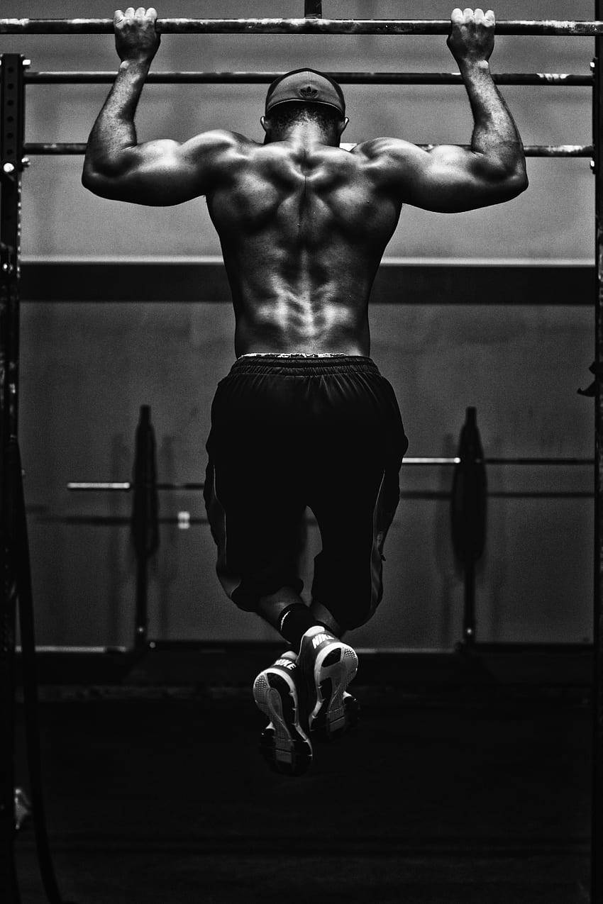 bodybuilders HD 桌布 壁紙 — 一個肌肉發達的男人掛在單色健身房的引體向上槓上，背部肌肉線條分明，專注而充滿力量。