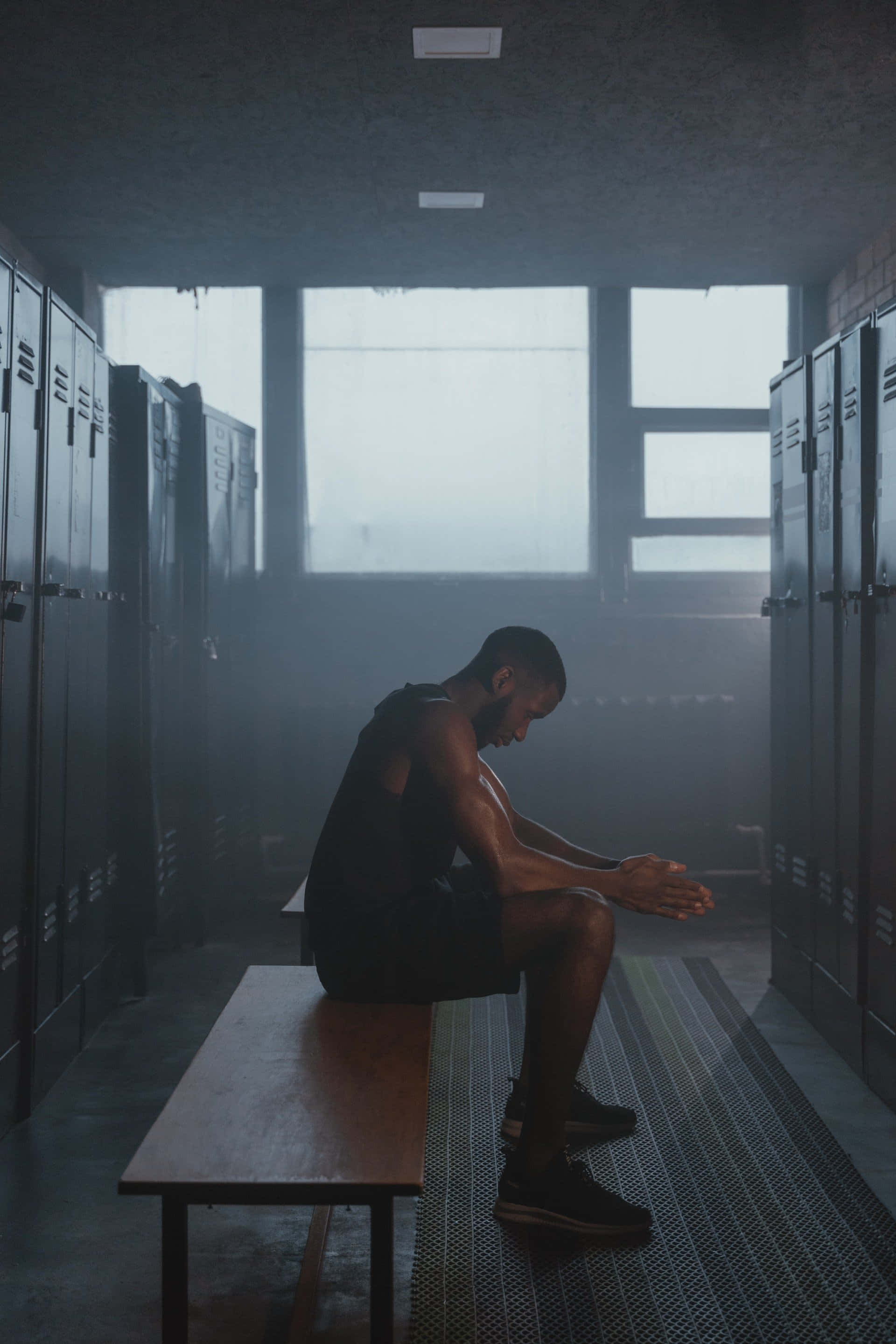 Lonely 桌布 壁紙 — 一個肌肉發達的男人獨自坐在昏暗更衣室的長凳上，雙手緊握，沐浴在霧氣窗戶透進的柔和光線中。