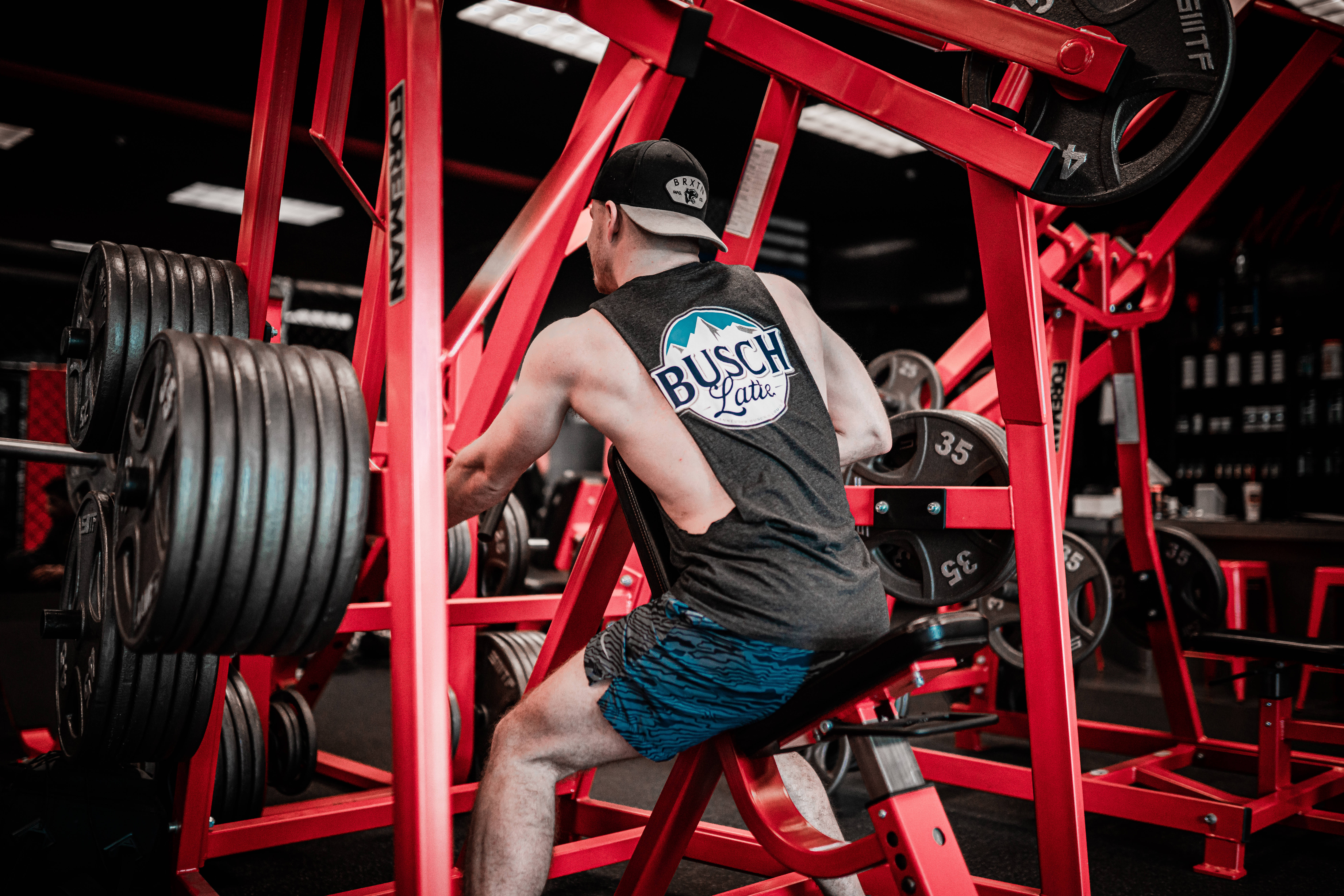 Weightlifting 桌布 壁紙 — 一個肌肉發達的男人穿著Busch Light背心，在明亮的螢光燈下於紅色健身器材上舉重。