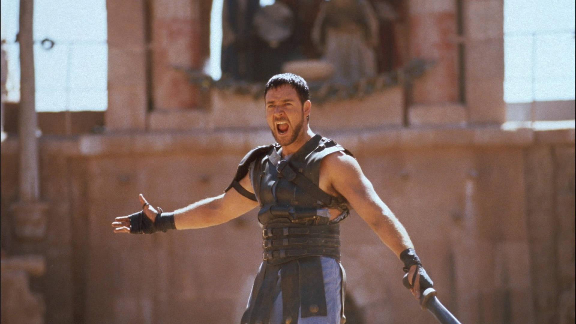 Russell Crowe 桌布 壁紙 — 一個肌肉發達的男人穿著古代盔甲，在陽光照耀的石頭競技場中張開雙臂咆哮。