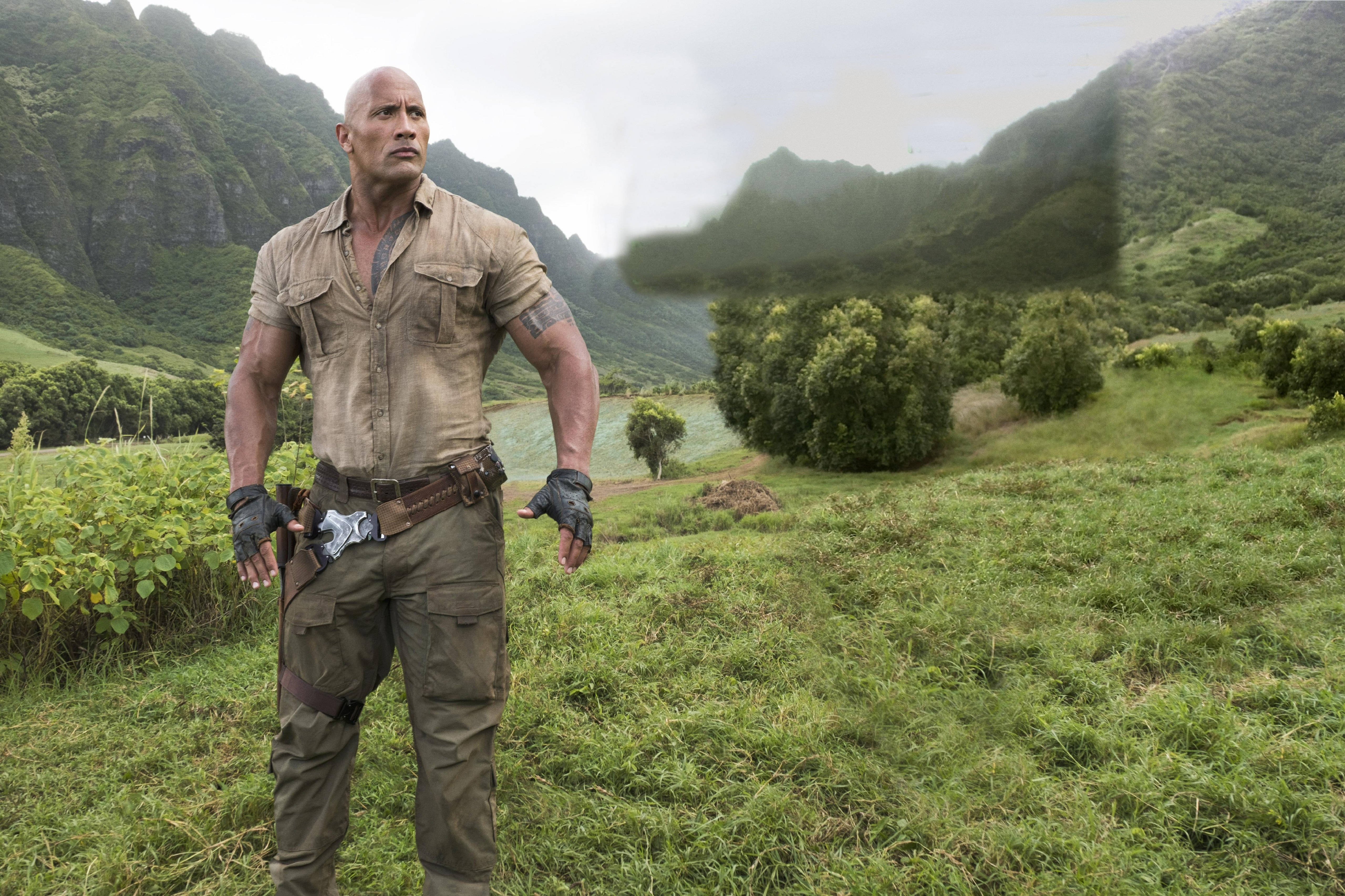 Dwayne 桌布 壁紙 — 一個肌肉發達的男人穿著堅固裝備，在一個被高聳霧氣繚繞的山脈包圍的茂密綠色山谷中警覺地站著。