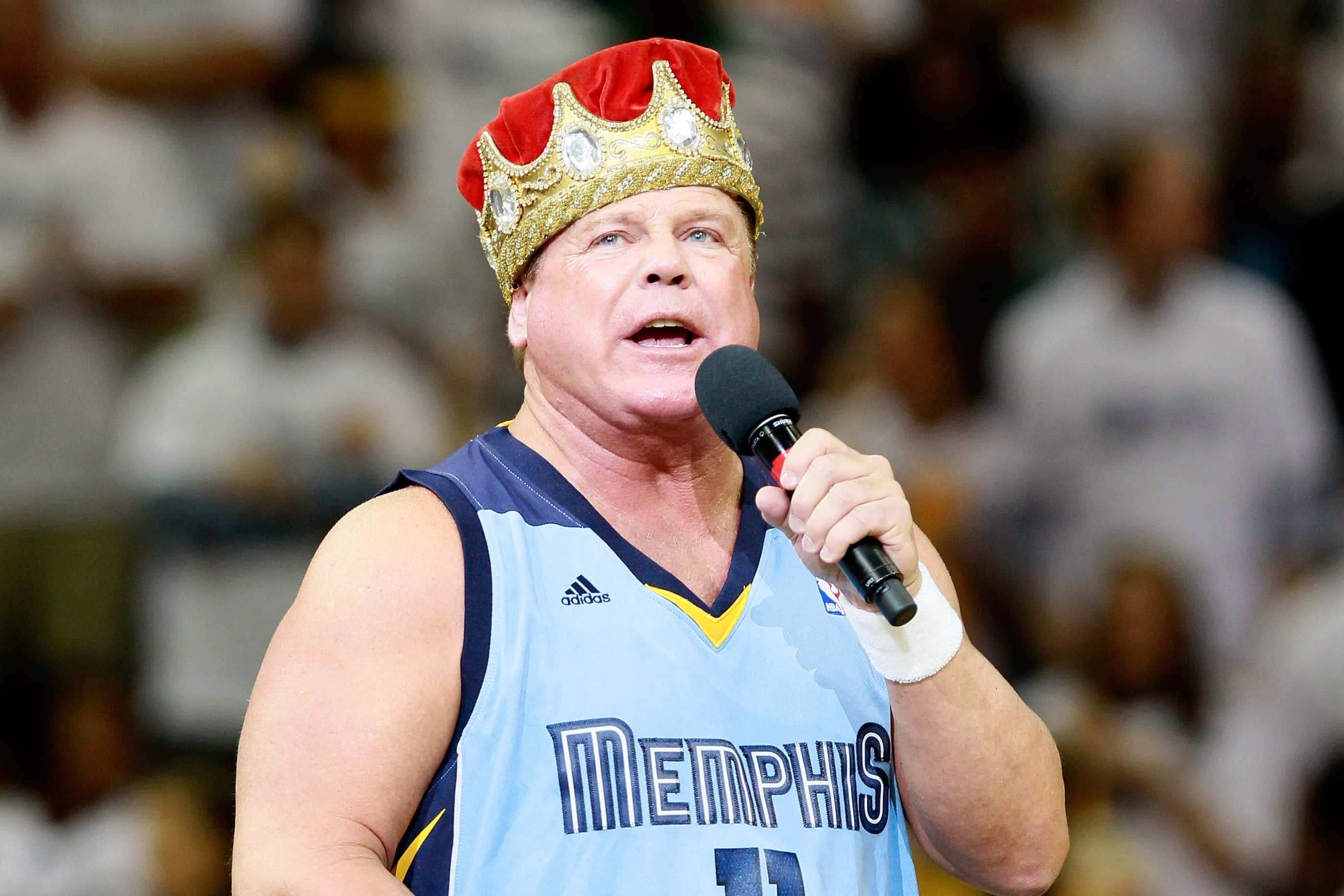 WWE 桌布 壁紙 — 一個肌肉發達的男人穿著曼菲斯球衣，戴著紅金皇冠，在籃球比賽中對著麥克風說話。