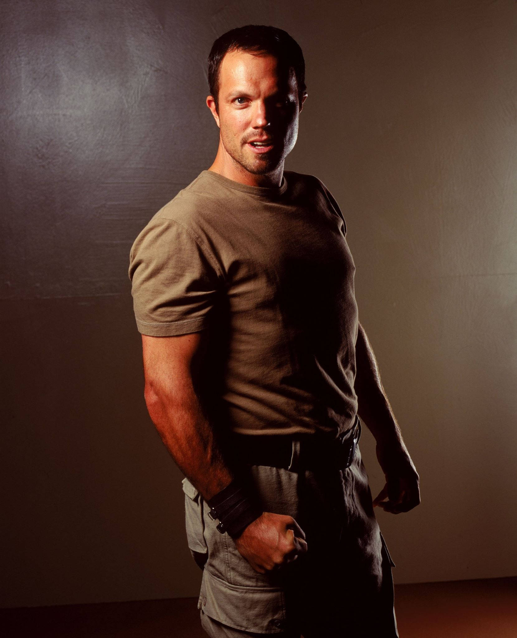 adam baldwin 桌布 壁紙 — 一個肌肉發達的男人穿著棕色T恤，自信地站在深色紋理牆前，燈光效果戲劇化。