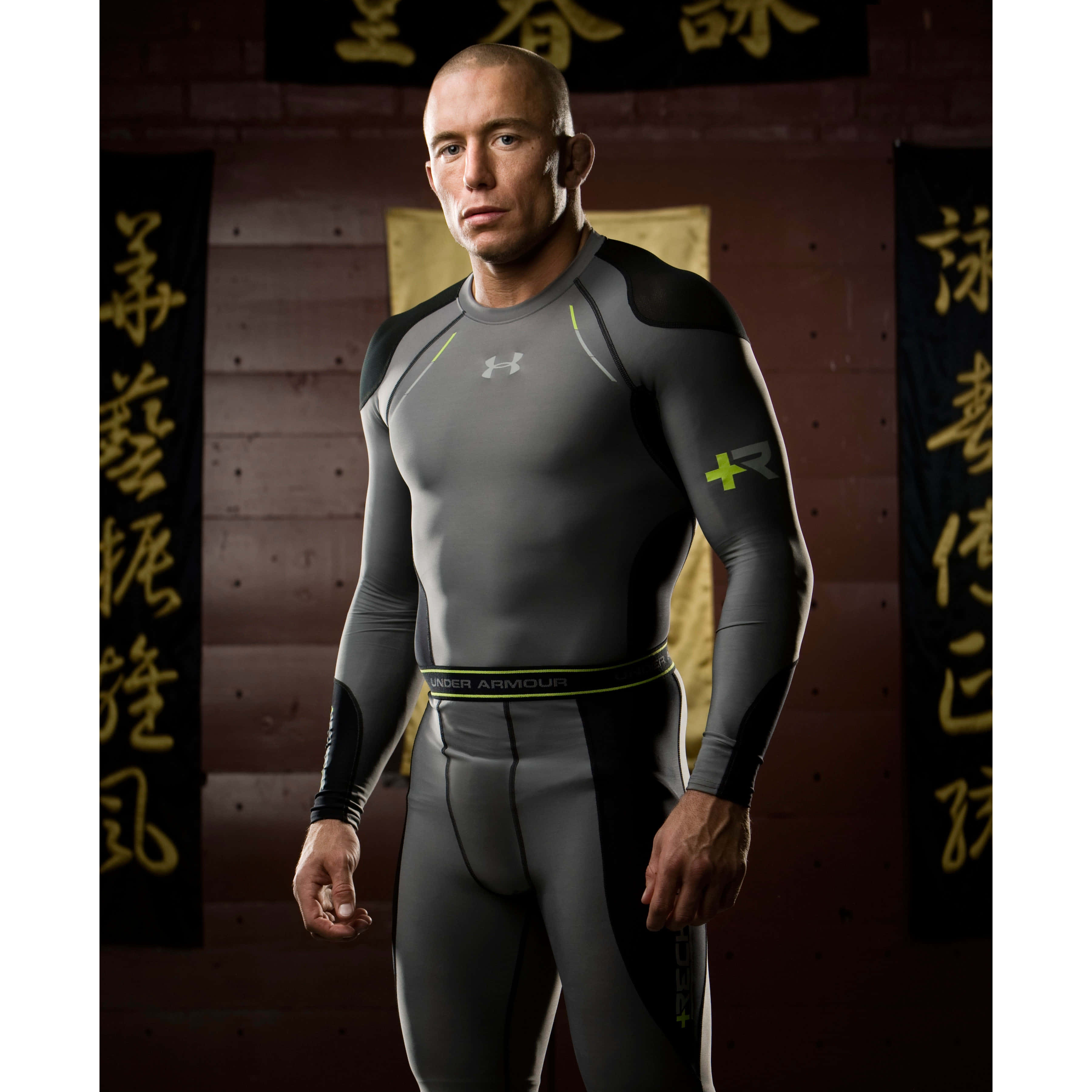 Under Armour 桌布 壁紙 — 一個肌肉發達的男人穿著灰色和黑色的Under Armour壓縮裝備，自信地站在一面裝飾著中國書法橫幅的磚牆前。