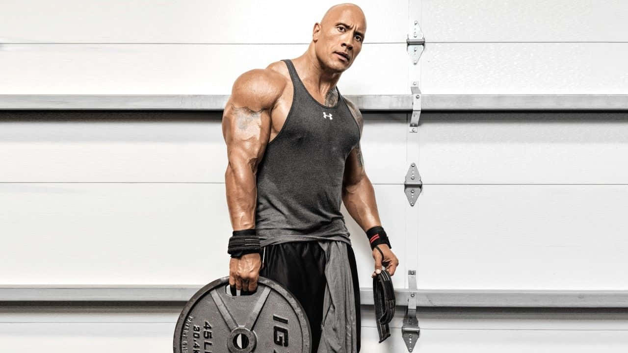The Rock 桌布 壁紙 — 一個肌肉發達的男人穿著灰色背心，在身後有白色捲簾門的車庫裡拿著一個沉重的槓鈴。