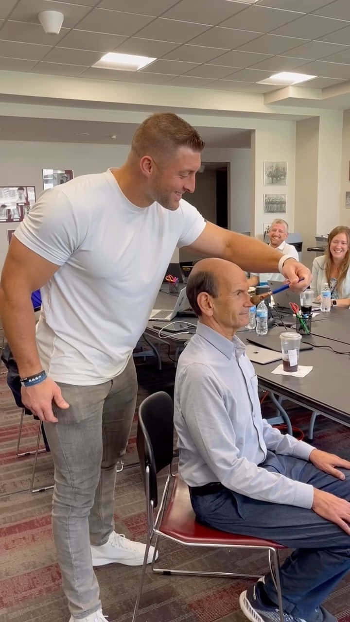 Tim Tebow 桌布 壁紙 — 一個肌肉發達的男人穿著白色T恤，在會議桌上戲弄地輕拍一位坐著的年長男人的頭，背景中其他人在微笑。