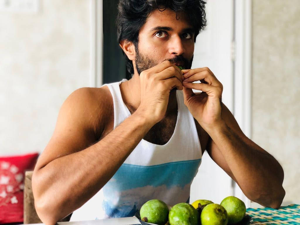 vijay devarakonda 4K 桌布 壁紙 — 一個肌肉發達的男人穿著白色背心，坐在桌子旁吃綠色水果，附近還有更多水果。