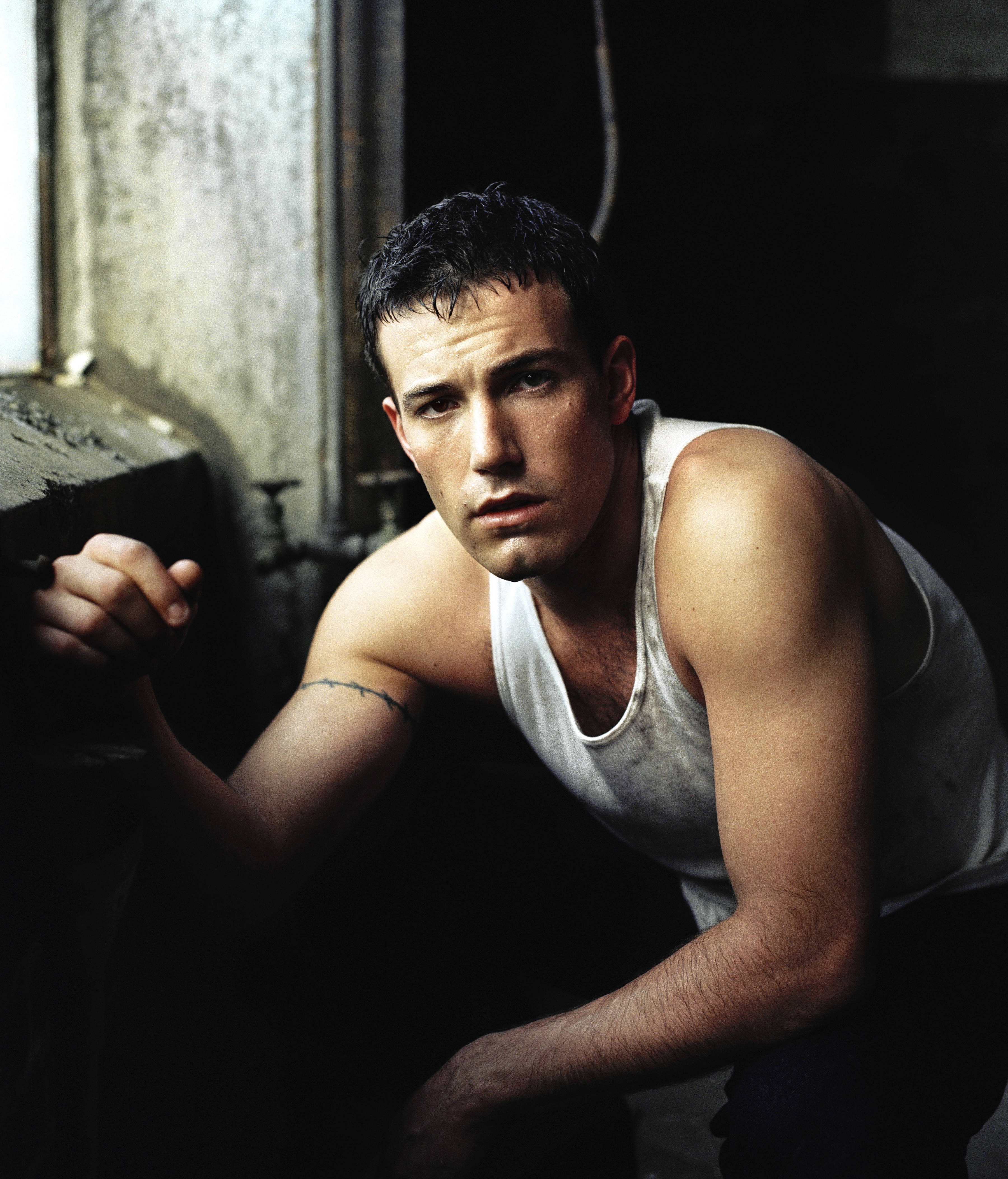 Ben Affleck 桌布 壁紙 — 一個肌肉發達的男人穿著白色背心，靠在昏暗工業空間的粗糙牆壁上，他銳利的目光直視鏡頭。