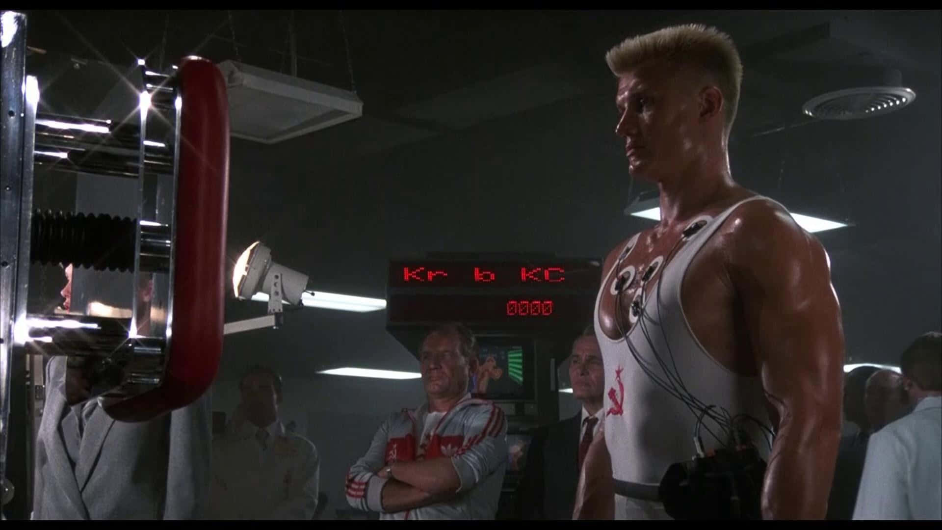 Dolph Lundgren 桌布 壁紙 — 一個肌肉發達的男人穿著白色背心，站在一個光線昏暗、有紅色器材和旁觀者注視他的健身房裡。