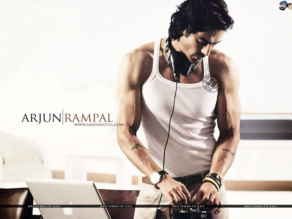 Arjun Rampal 桌布 壁紙 — 一個肌肉發達的男人穿著白色背心和耳機，在明亮簡約的房間裡俯身於筆記型電腦上。