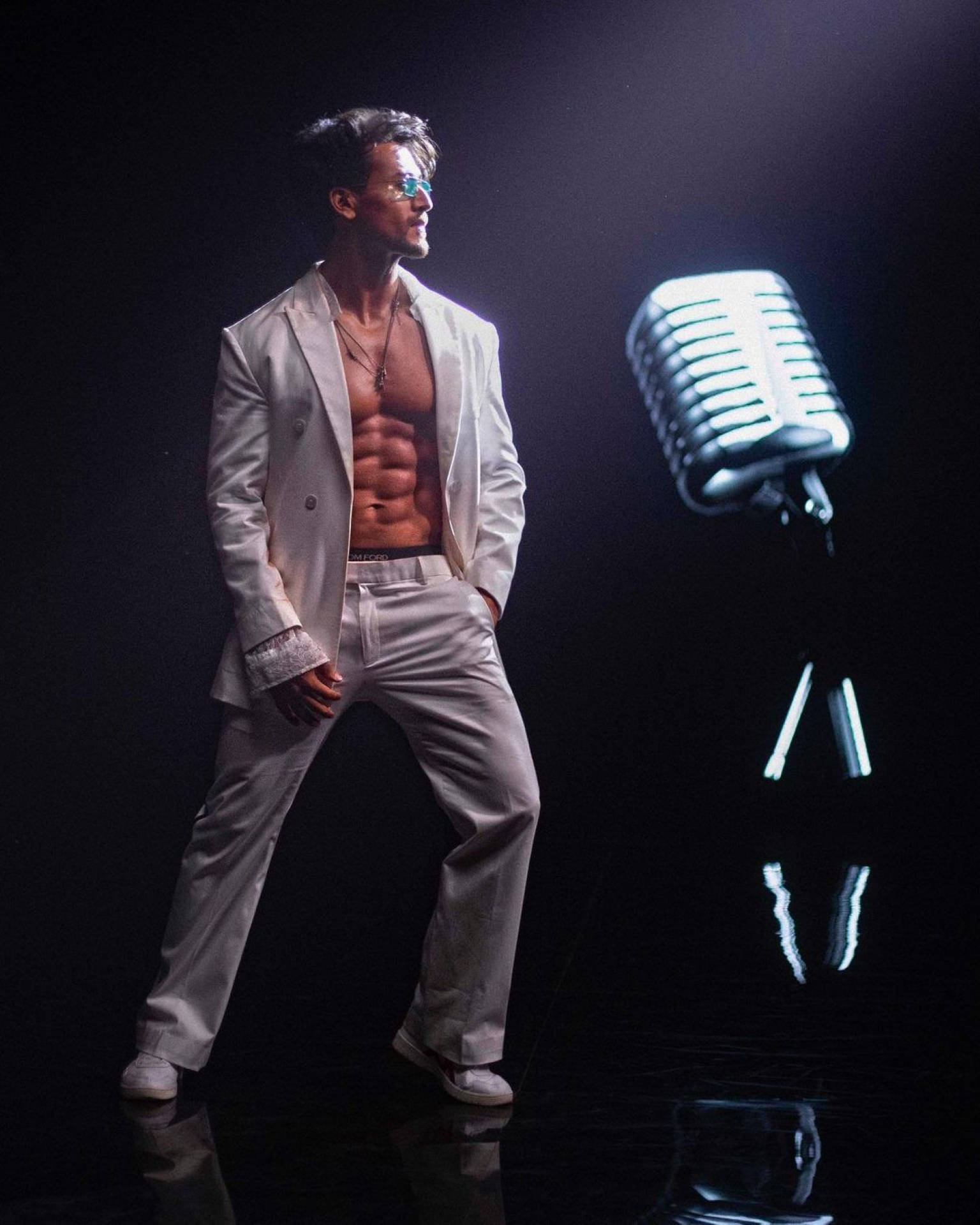 Tiger Shroff 桌布 壁紙 — 一個肌肉發達的男人穿著白色西裝，自信地站在黑暗反光舞台上一個發光的復古麥克風旁邊。