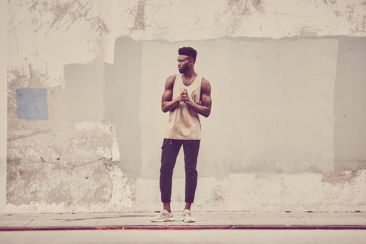 Jaylen Brown 桌布 壁紙 — 一個肌肉發達的男人穿著米色背心，雙手交握，自信地站在風化的城市牆前。