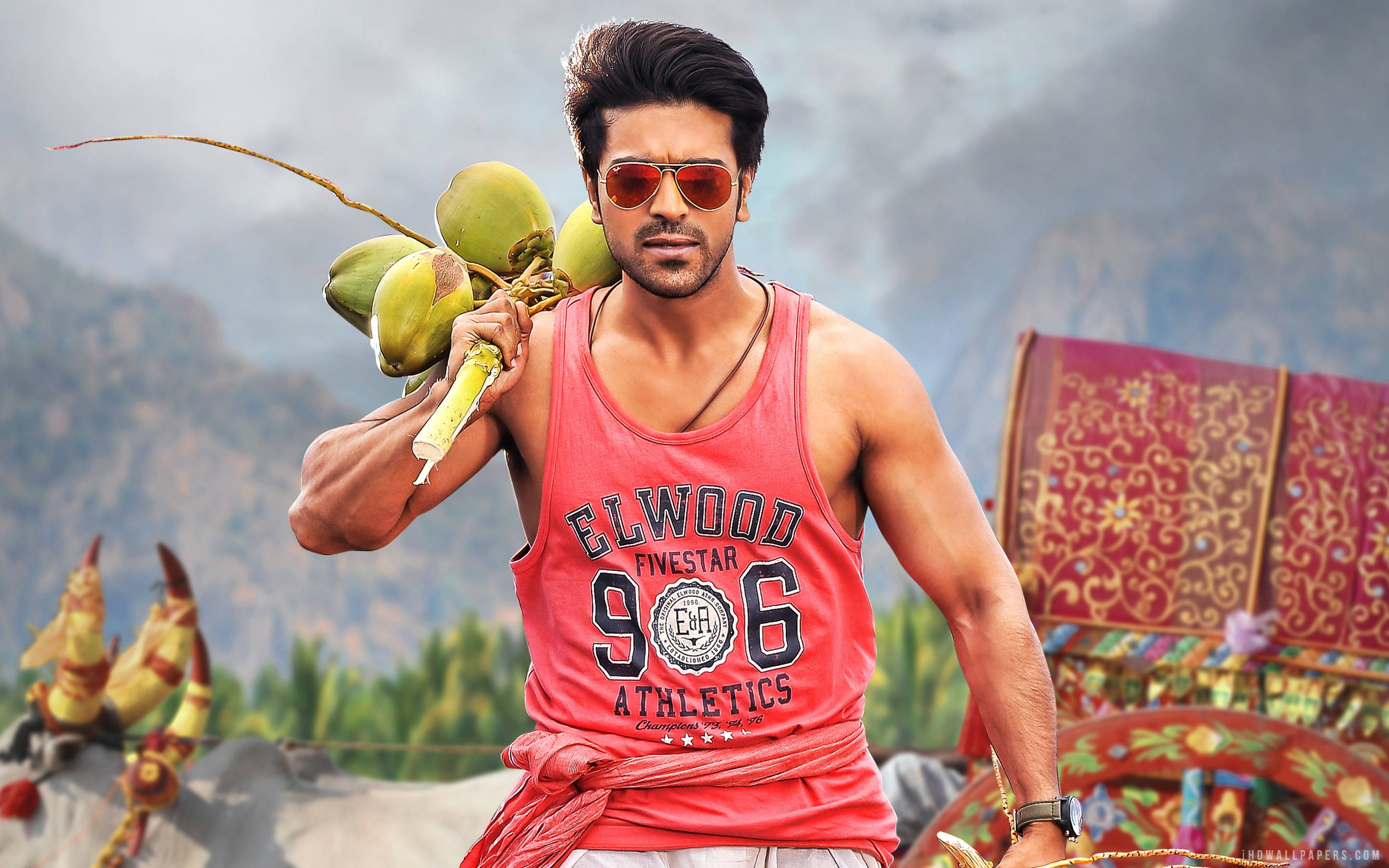 Ram Charan 桌布 壁紙 — 一個肌肉發達的男人穿著紅色背心和太陽眼鏡，在群山背景下肩上扛著綠色椰子。
