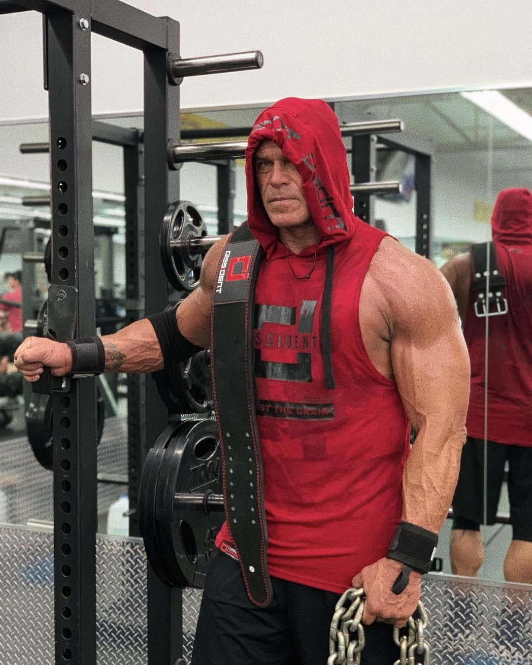 WWE 桌布 壁紙 — 一個肌肉發達的男人穿著紅色連帽衫和舉重腰帶，在健身房裡激烈地站著，手持鐵鏈並抓住深蹲架。